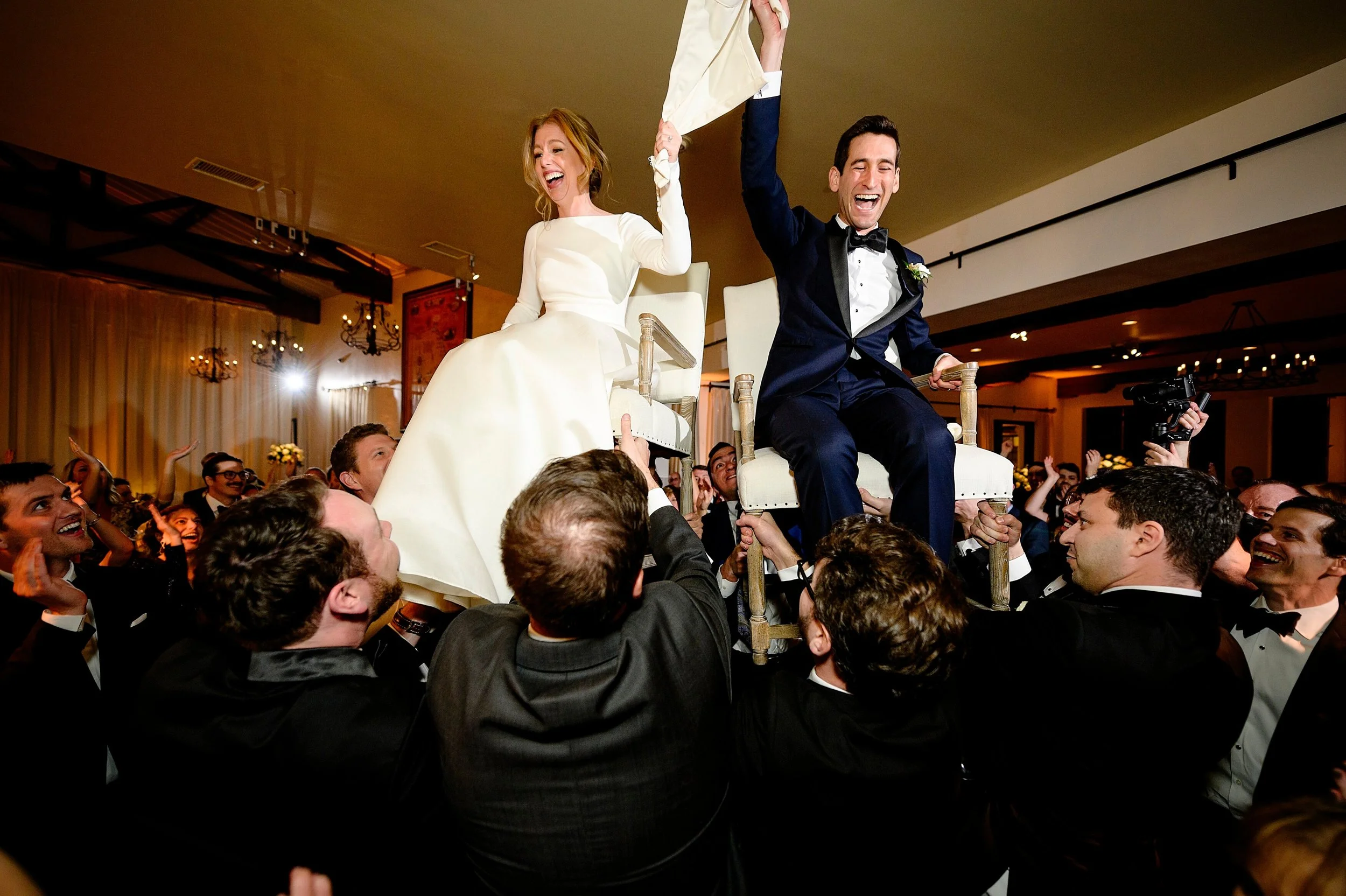 A bride and groom sitting on chairs elevated above a crowd, celebrating at their wedding reception. Both are smiling and holding hands in the air.