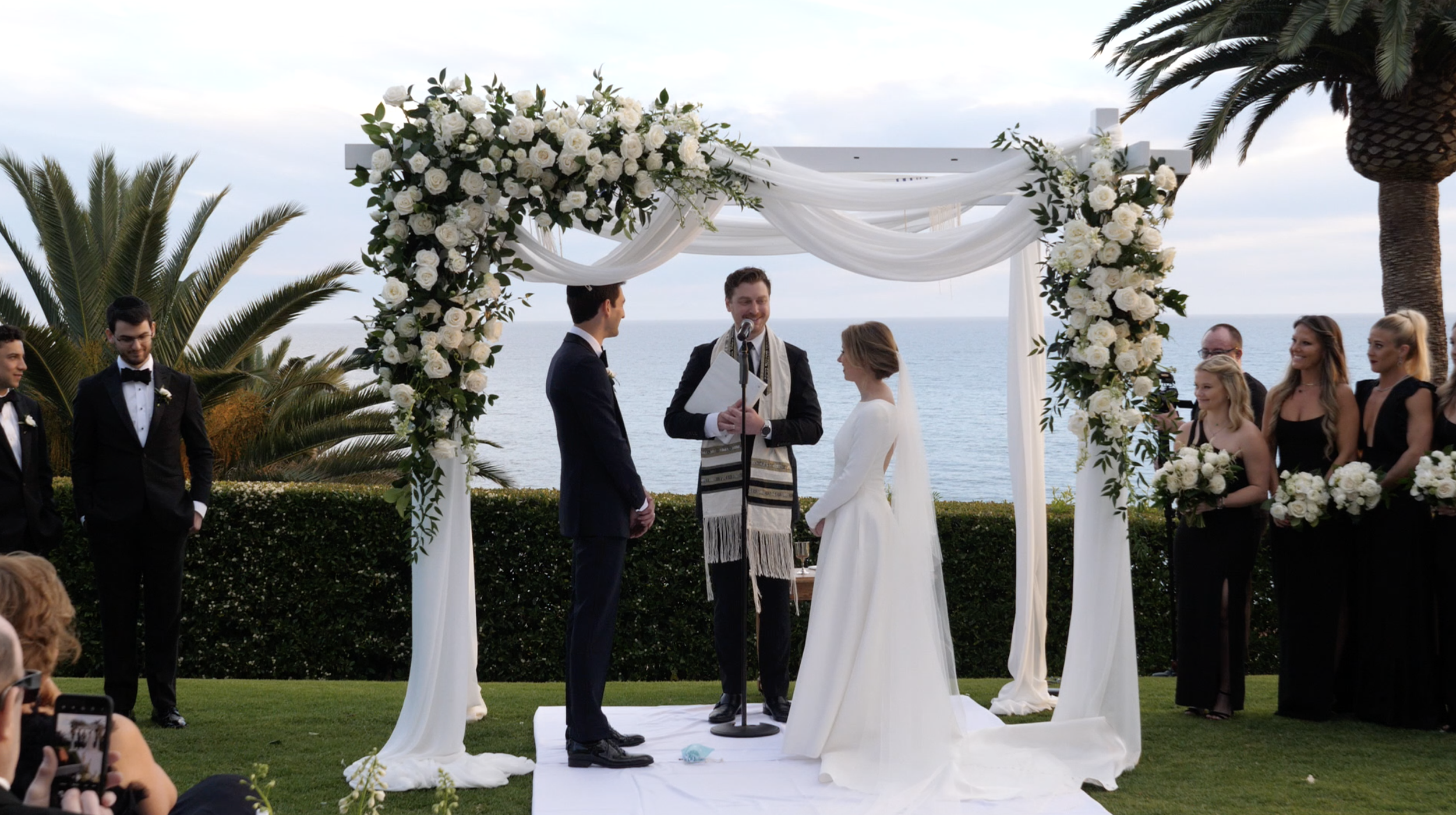 A wedding ceremony taking place outdoors by the ocean, with a floral arch and a bride and groom standing before an officiant, while bridesmaids and groomsmen stand on either side, all dressed in formal attire.