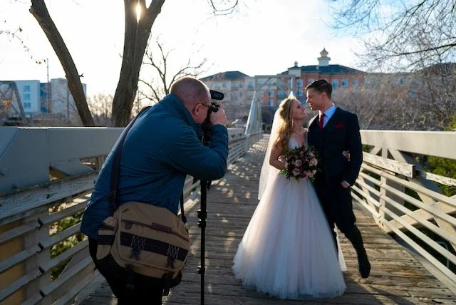 A wedding photoshoot on a bridge featuring a bride and groom posing with a videographer taking their picture.