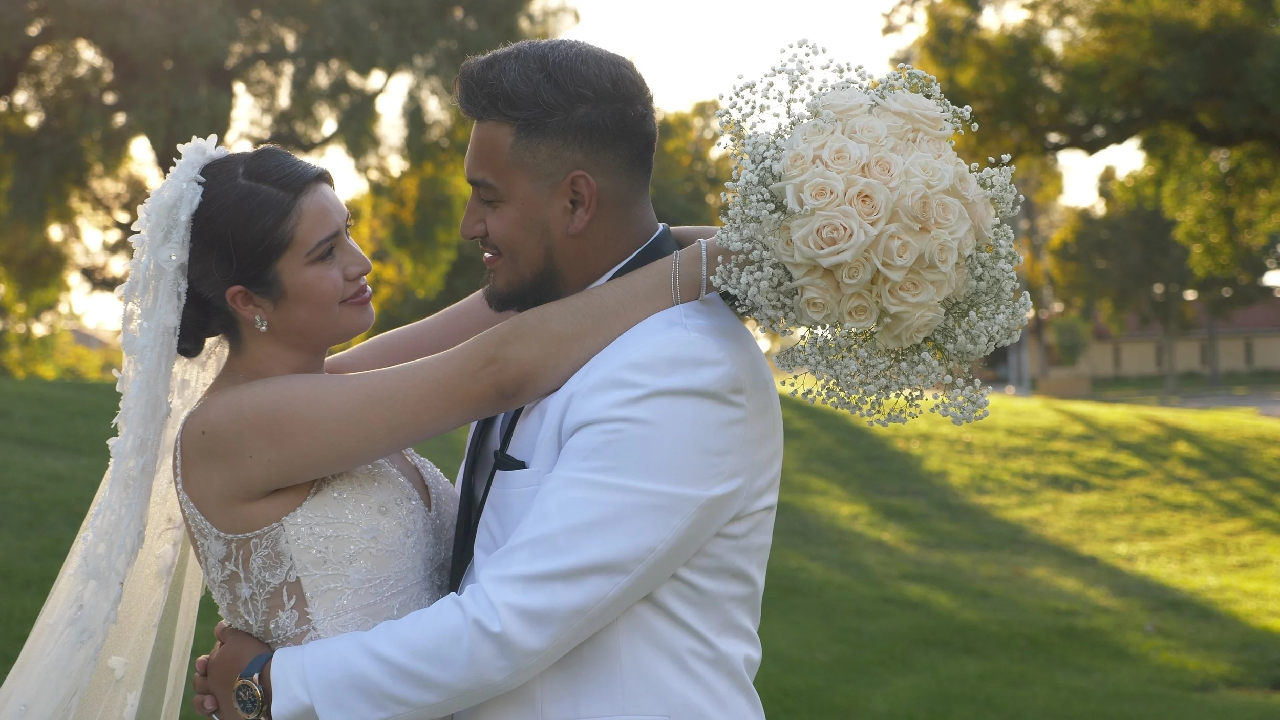 A bride and groom embrace outdoors during sunset, with the bride holding a bouquet of white roses and baby's breath. They look into each other's eyes with a background of green trees and sunlight.