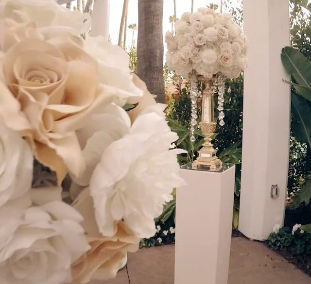 A tall floral arrangement with white and cream roses on a golden pedestal, surrounded by lush green foliage, placed on a white stand at an outdoor event.