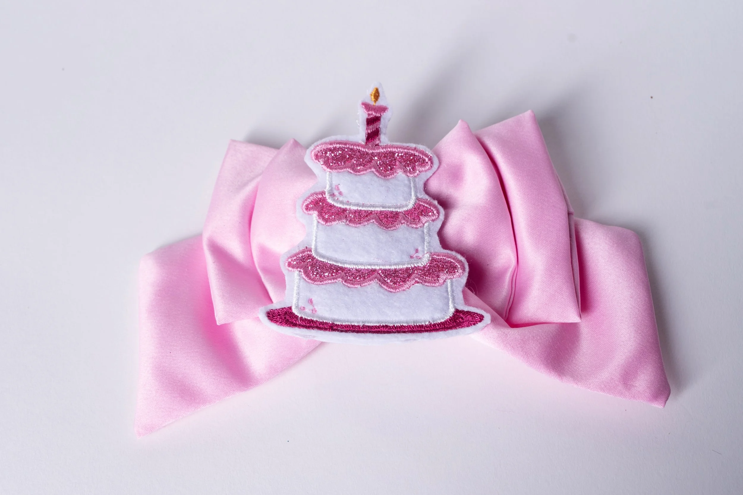Pink hair accessory with a glittery embroidered birthday cake with three tiers and a candle on top, attached to a pink satin ribbon.