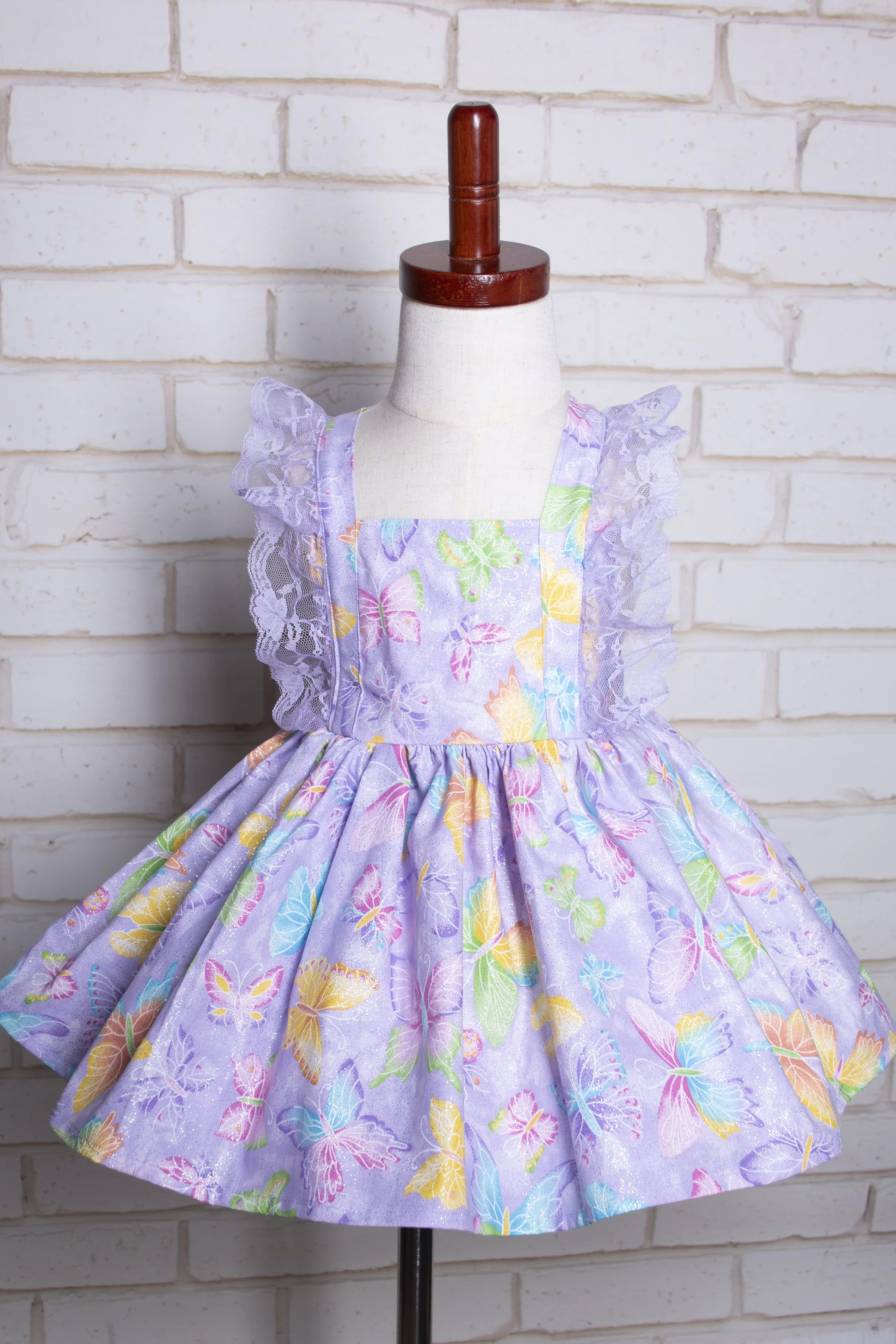 A lavender dress with butterfly and floral print on a dress form against a white brick wall.