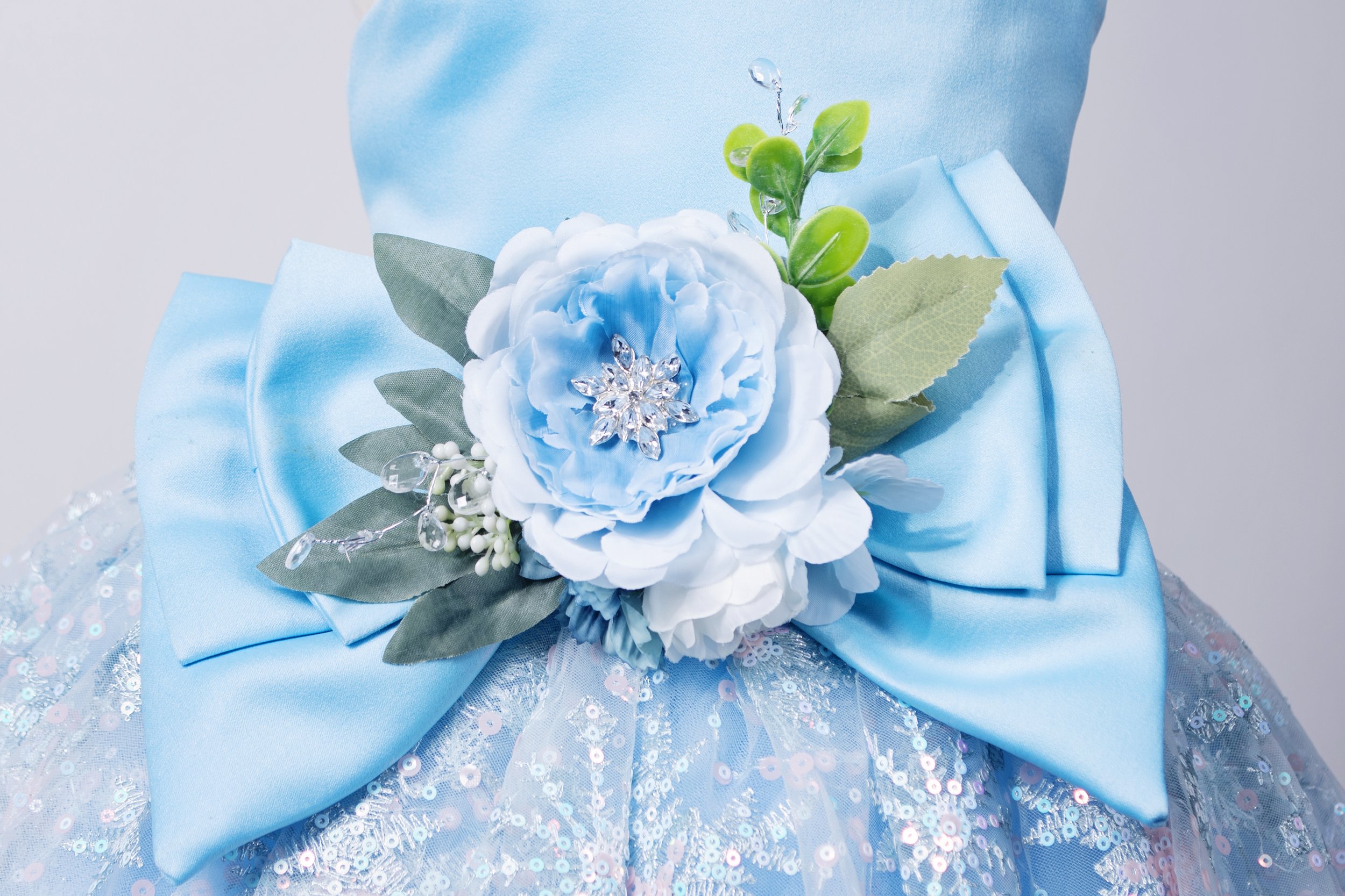 A close-up of a light blue satin fabric with a large satin bow and a floral embellishment consisting of a large light blue peony, greenery, and a crystal brooch, on a sequin-embellished fabric background.