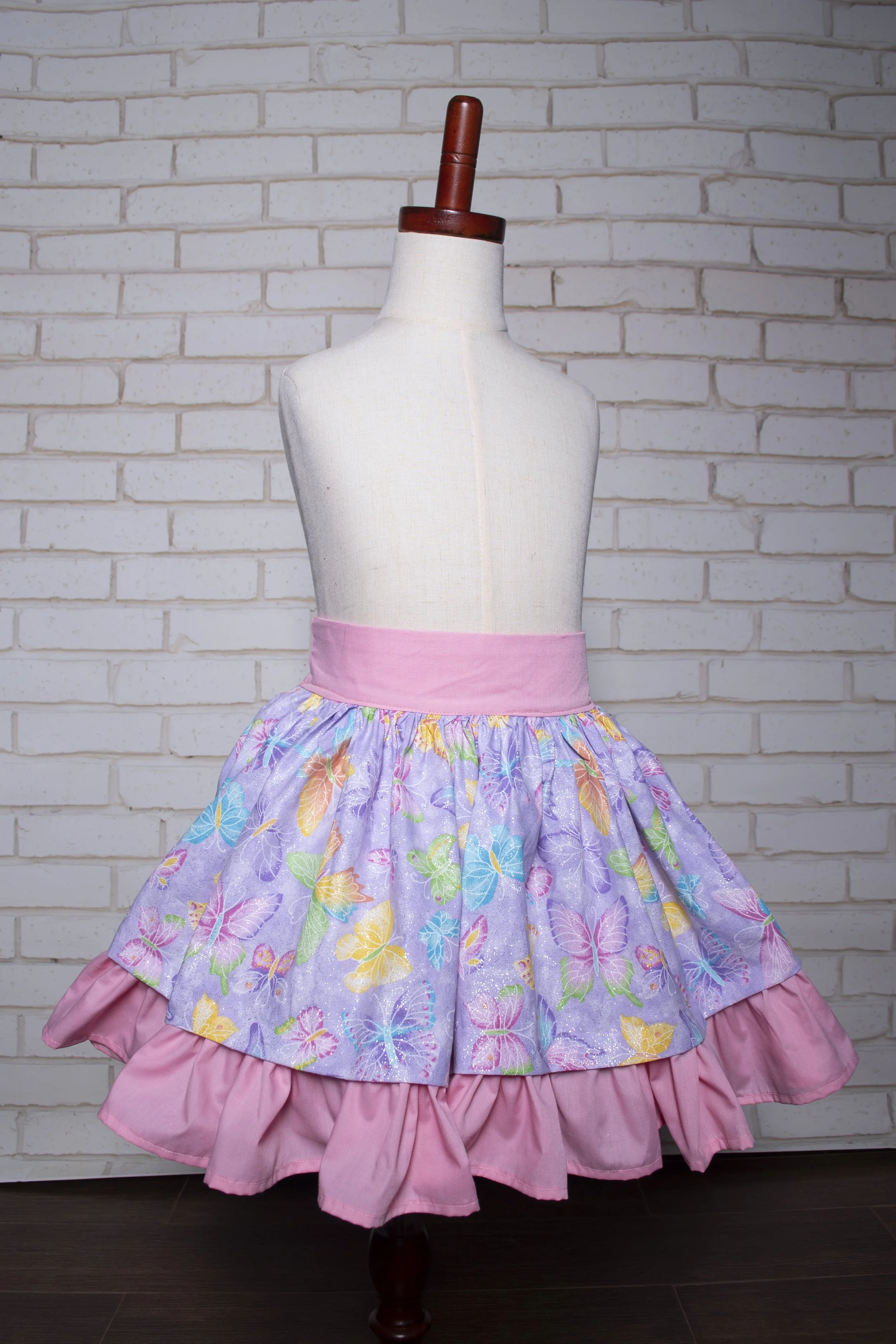 A dress with a pink waistband and a purple skirt featuring colorful butterfly patterns, displayed on a mannequin against a white brick wall.
