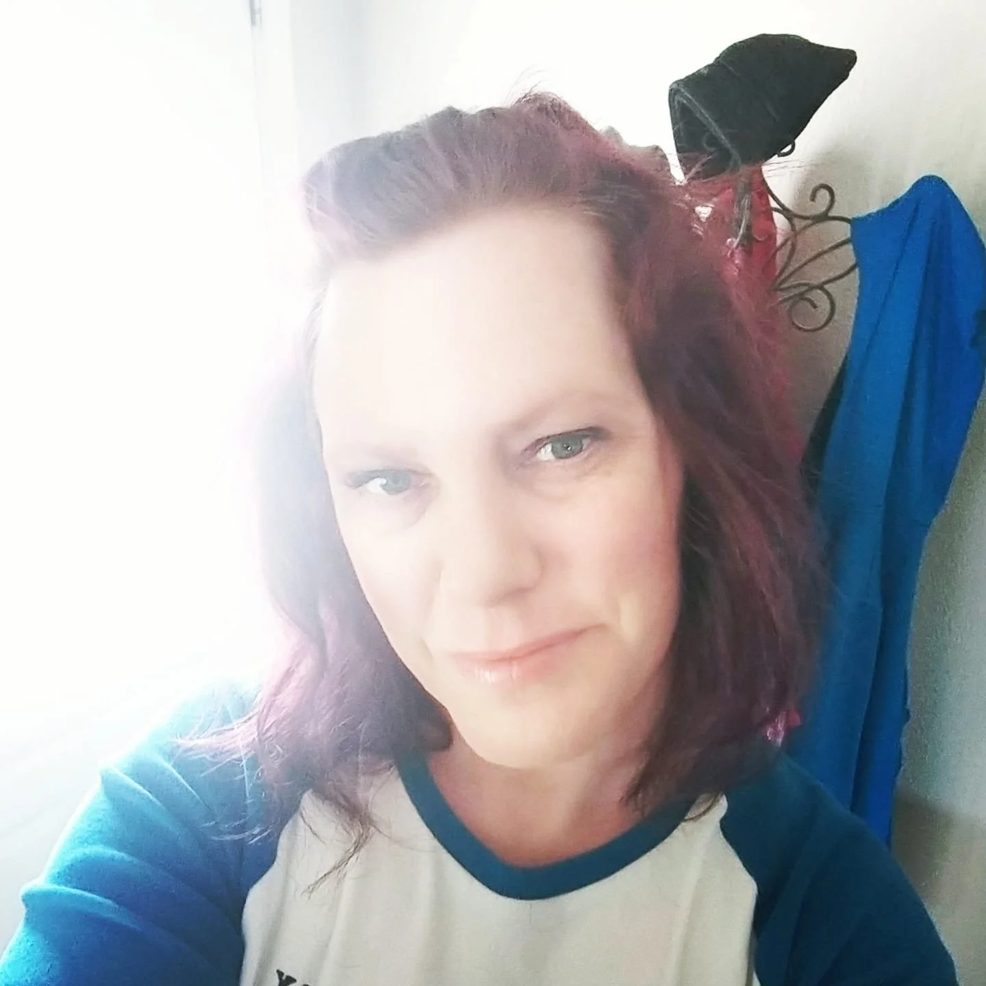 A woman with light-colored eyes and shoulder-length wavy red hair taking a selfie indoors near a window with sunlight streaming in.