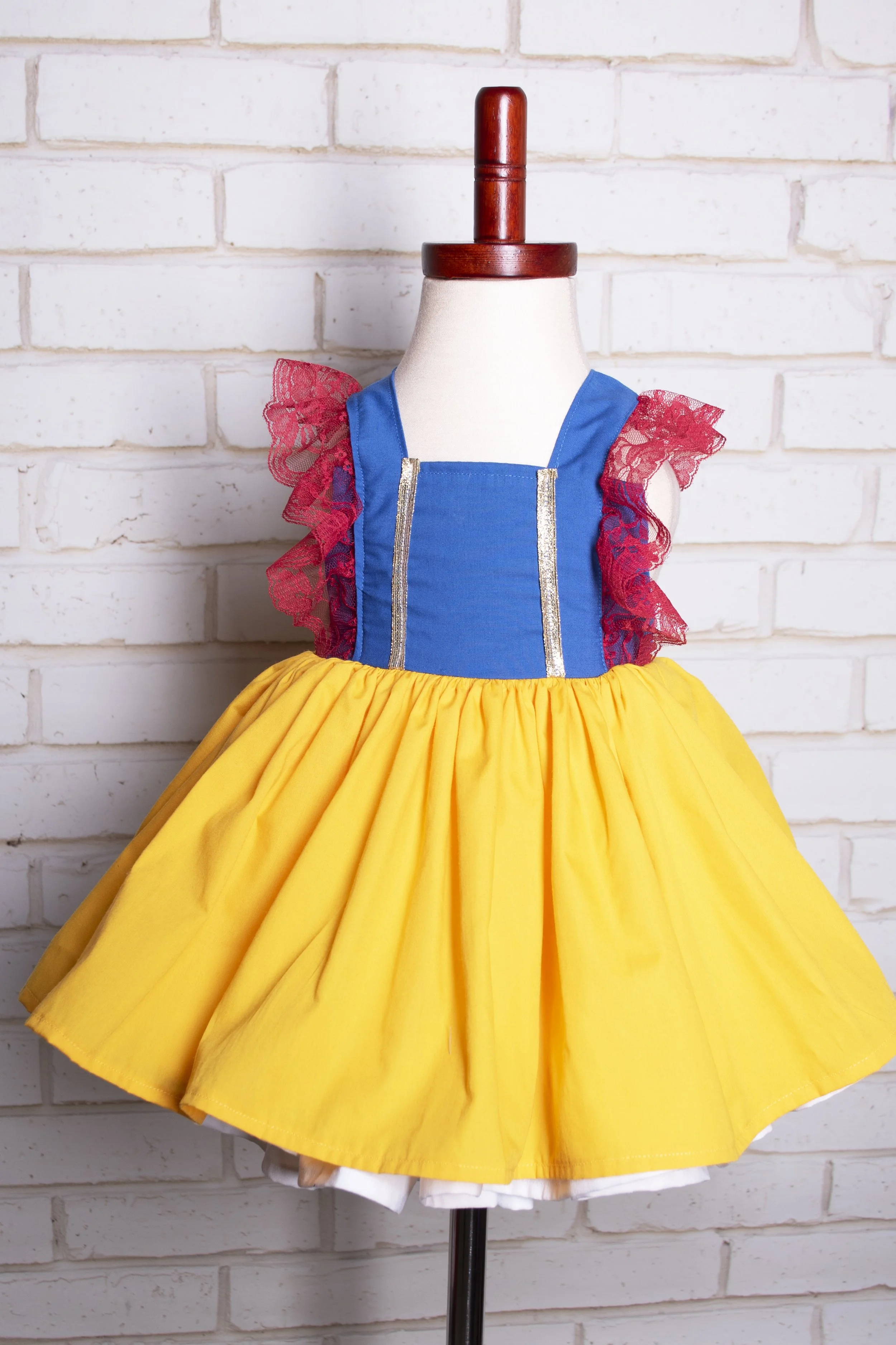 Children's dress designed to resemble Snow White, with a blue bodice, red lace sleeves, and a yellow skirt, displayed on a mannequin against a white brick wall.