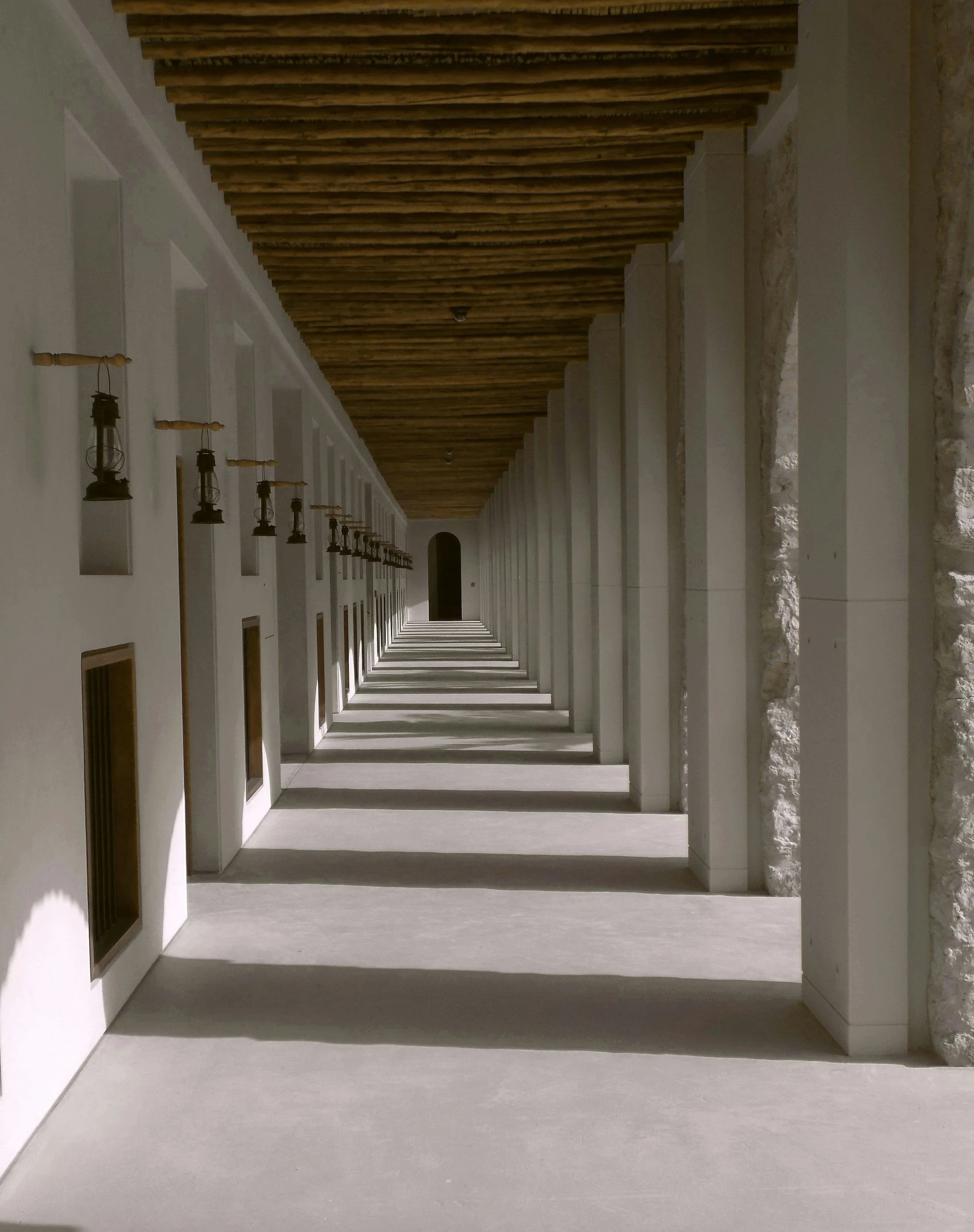 Long covered walkway with white walls, archways, lantern-style lights on the left, and a wooden ceiling.