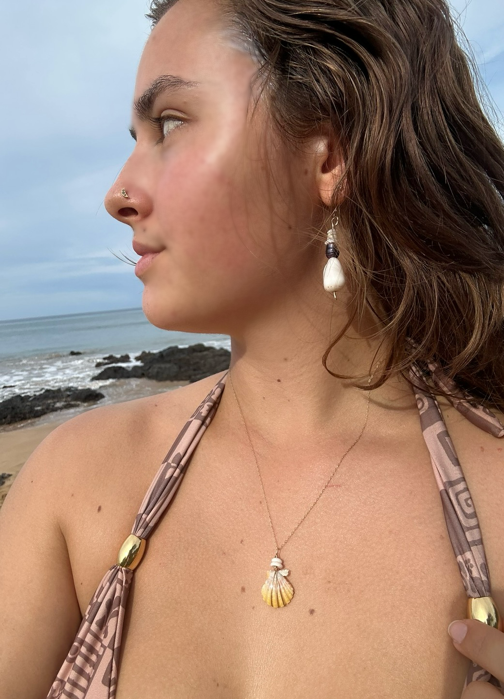 A woman with brown hair wearing shell jewelry at the beach with ocean and rocks in the background.