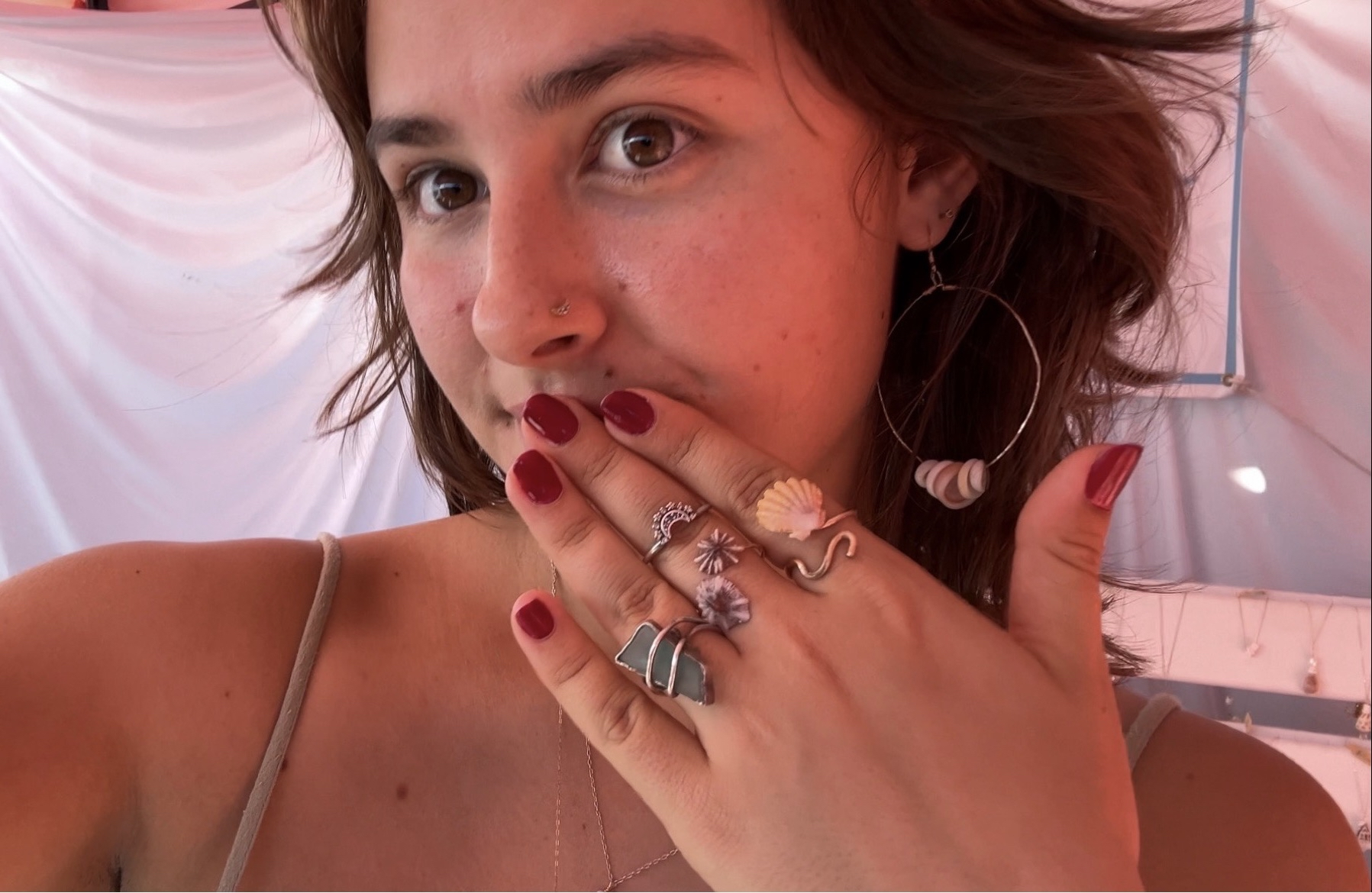 A woman with wavy brown hair and brown eyes, wearing large hoop earrings and a nose ring, holds her hand up to her mouth showing multiple rings and red nail polish. She is wearing a thin-strapped top and a delicate necklace. The background is a pastel-colored tent interior.