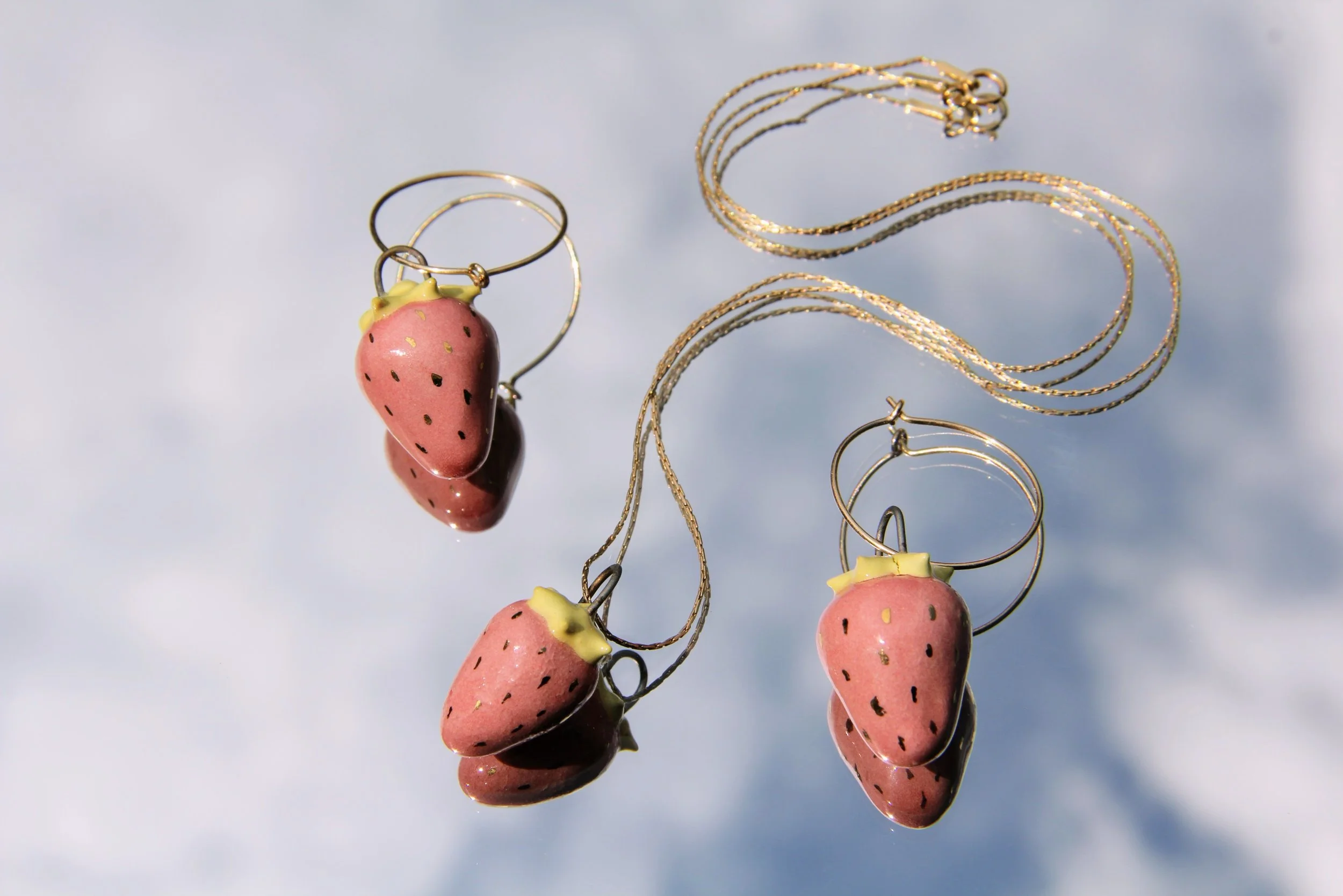 Pink strawberry-shaped earrings next to gold chain necklaces against a light blue background.