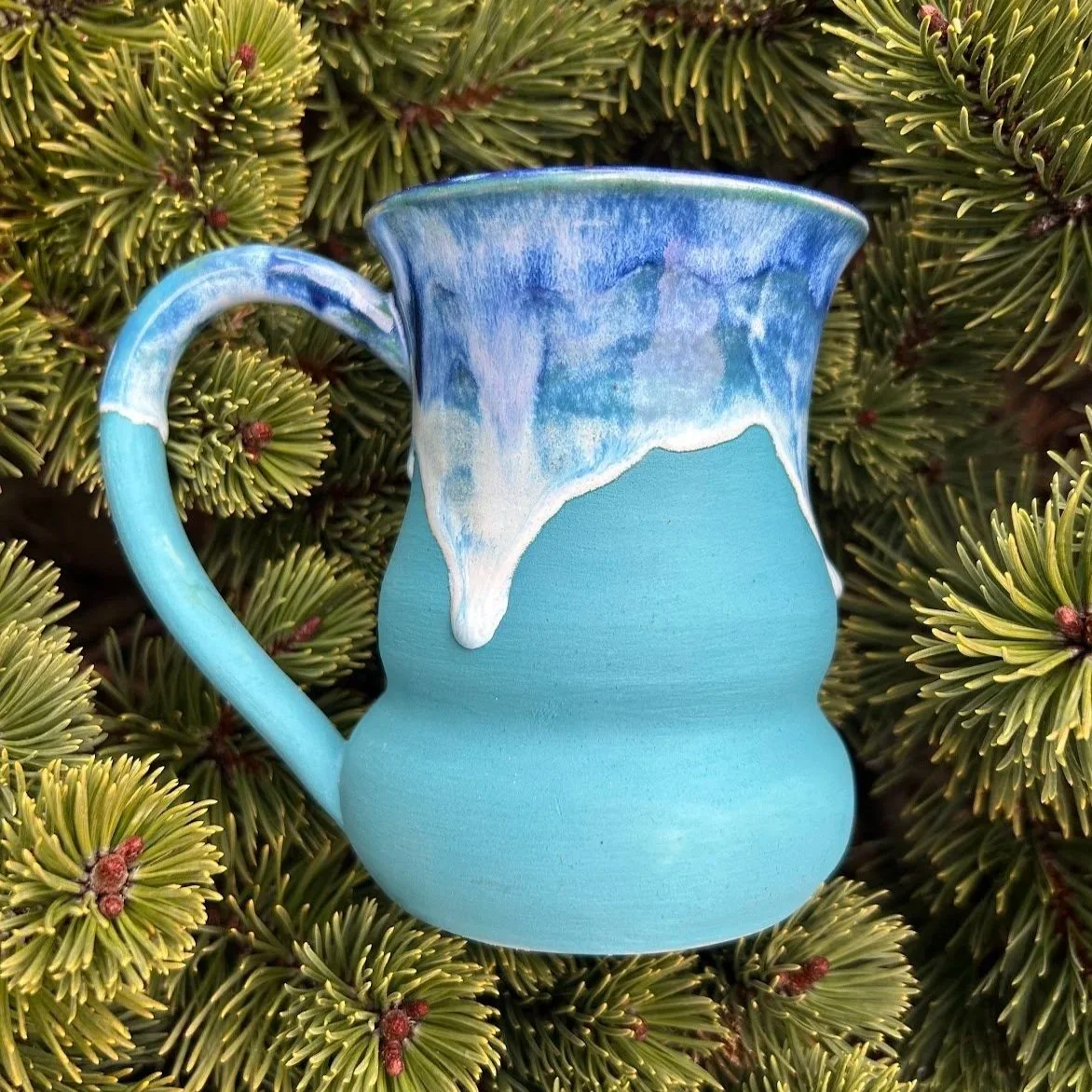Ceramic pitcher with a blue and white glaze drip design, placed on green pine branches.
