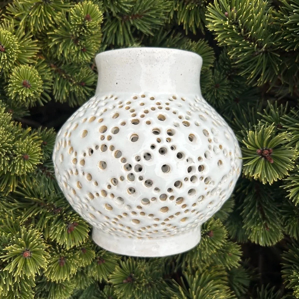 White ceramic vase with perforated, honeycomb-like design, surrounded by green pine branches.