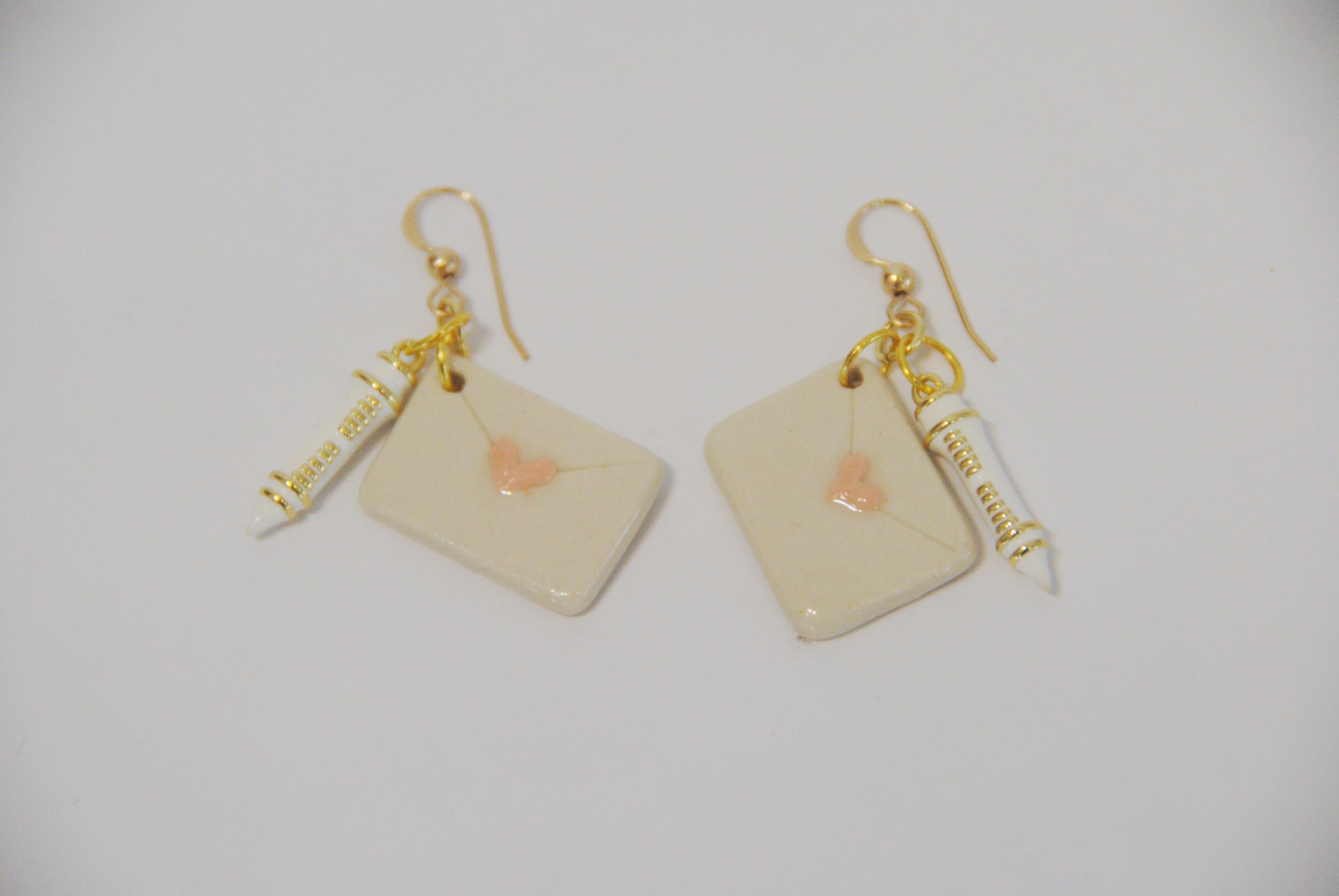 Pair of earrings with miniature clay items resembling a lipstick and a square card with a pink heart, connected by gold hooks on a white background.