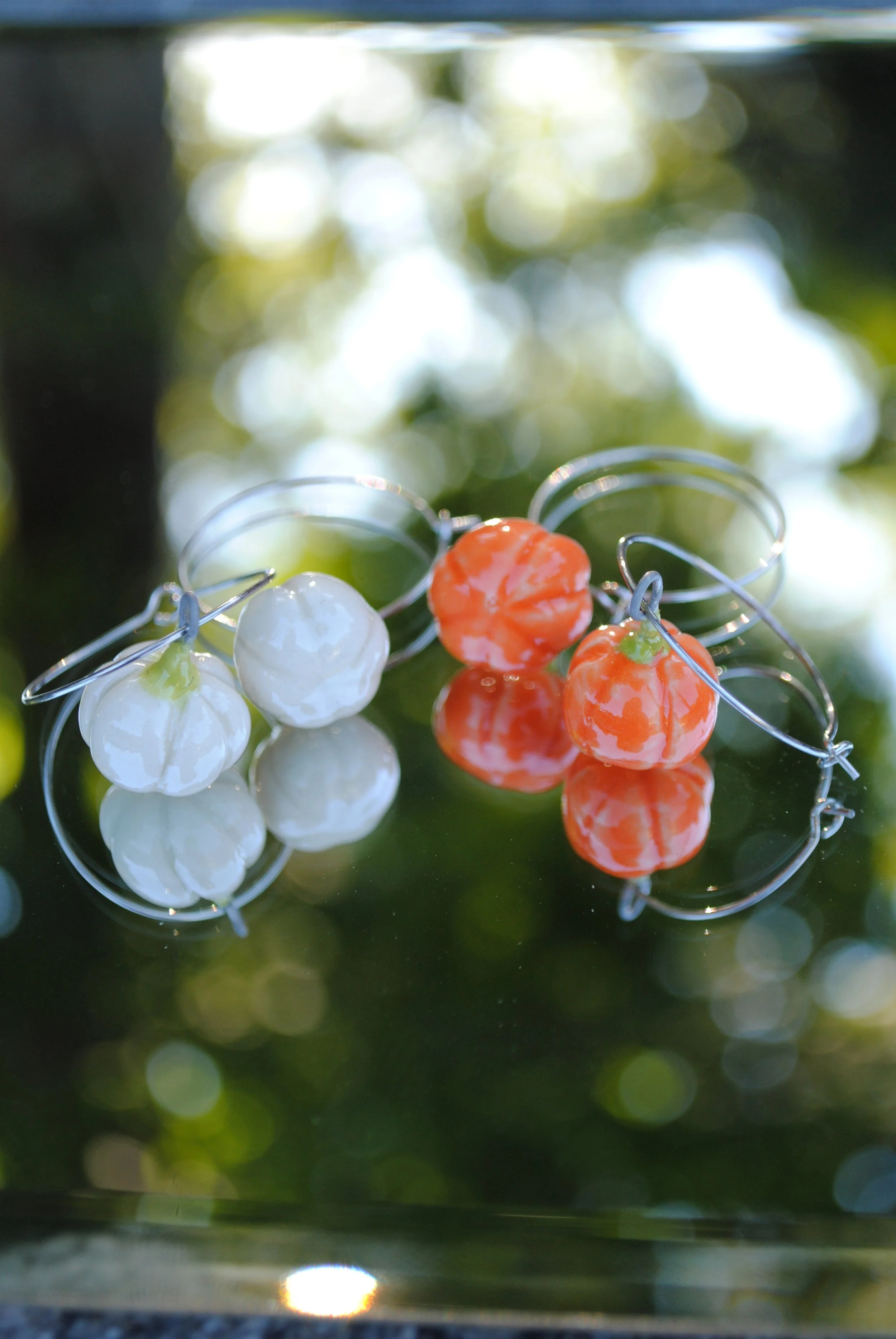 Close-up of pearl and orange jewelry with floral accents, placed on a mirror reflecting the jewelry, with a bokeh background of sunlight filtering through greenery.