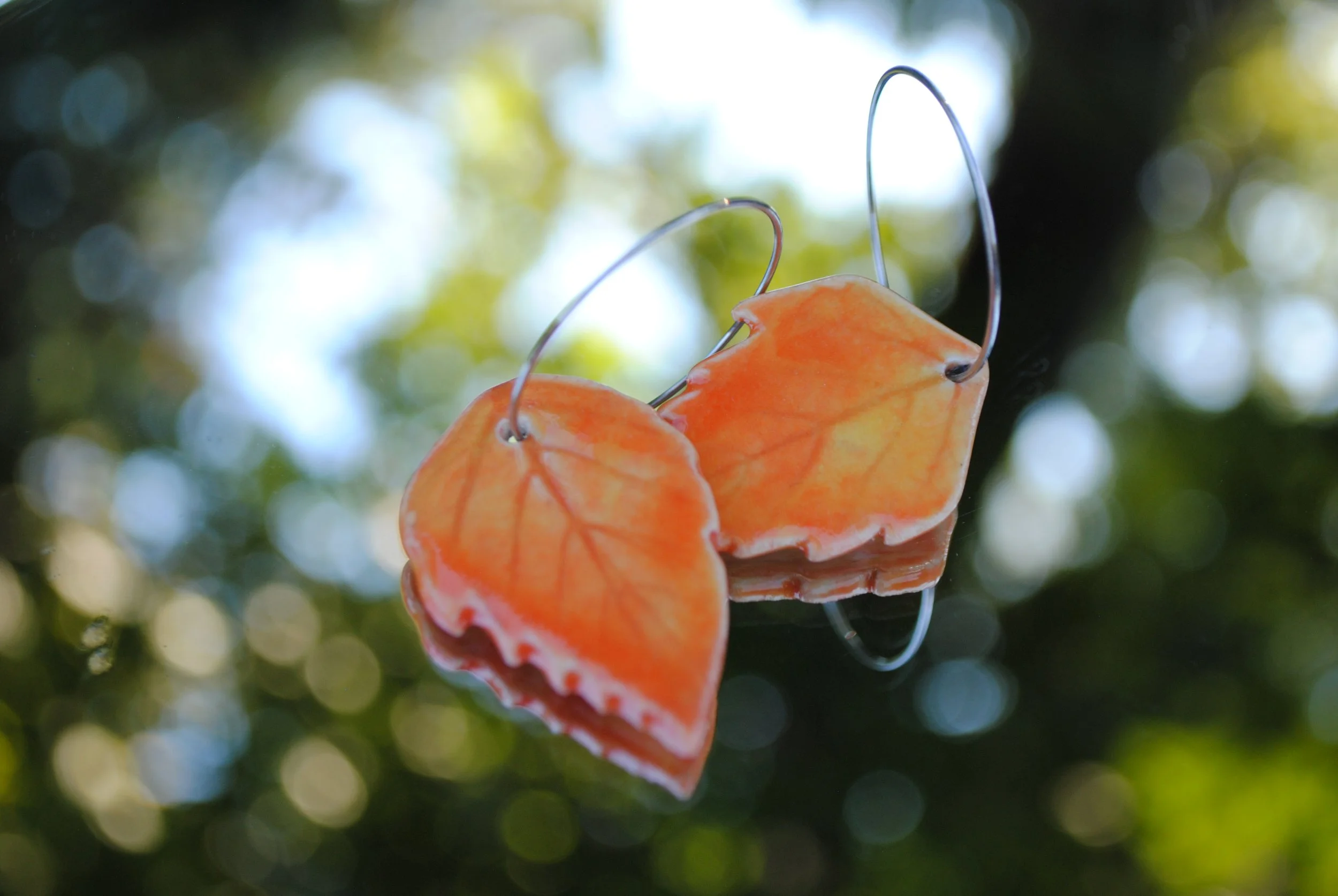 Orange butterfly-shaped earrings hanging against a blurred background of green and blue foliage with sunlight filtering through.