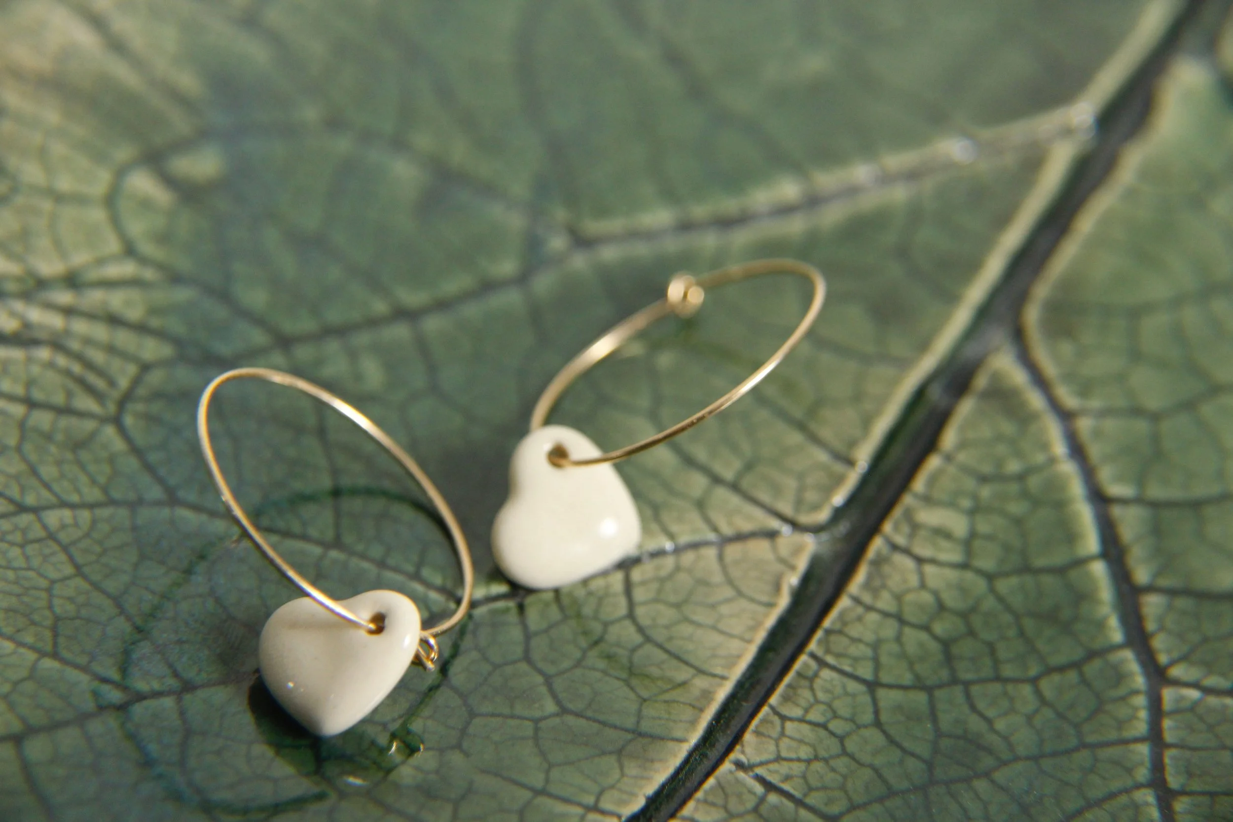 A pair of delicate earrings with white, heart-shaped beads and thin gold hoops lie on a textured green leaf.