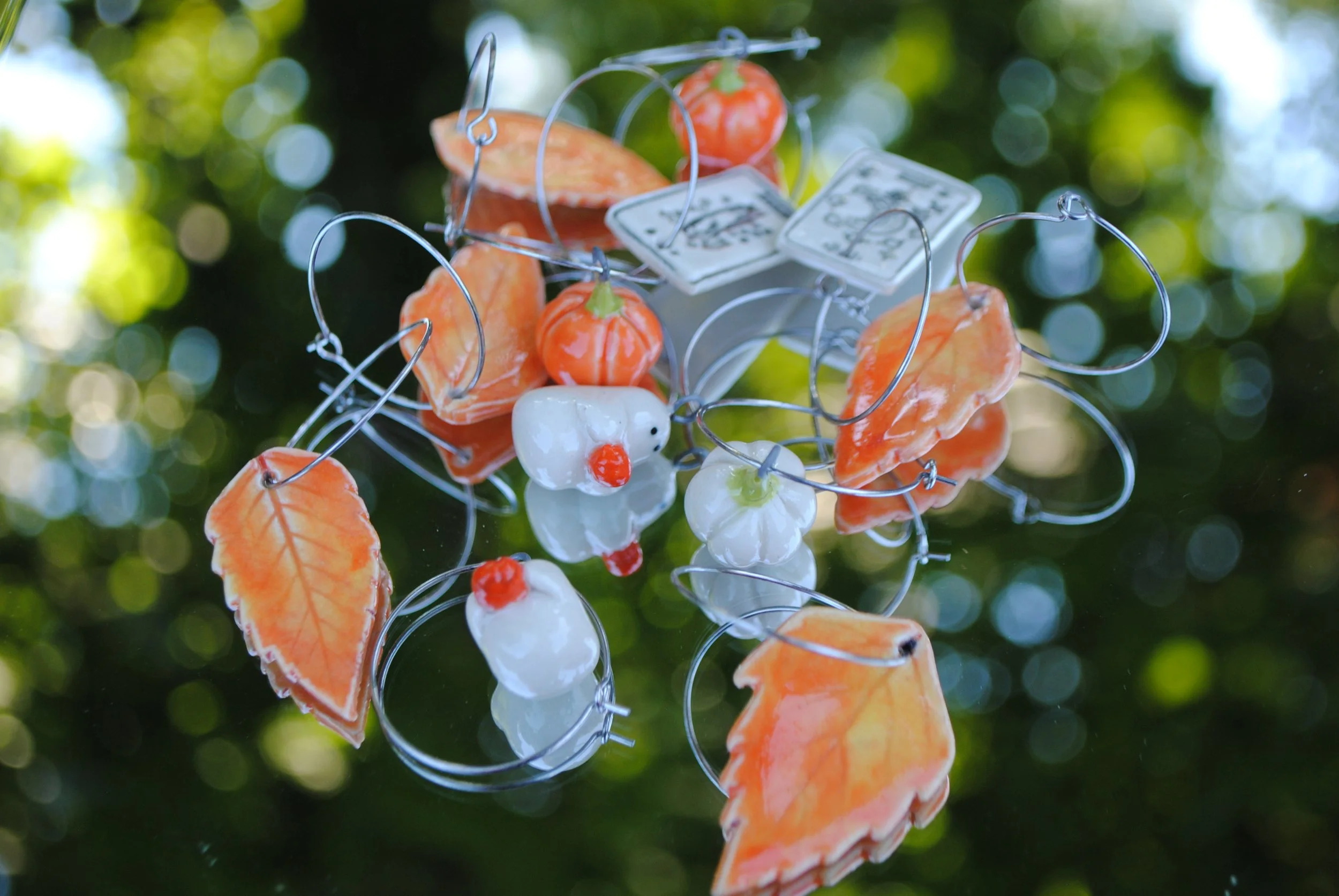 Glass art hanging outdoors featuring pumpkins, leaves, and a snowman, with green bokeh background.