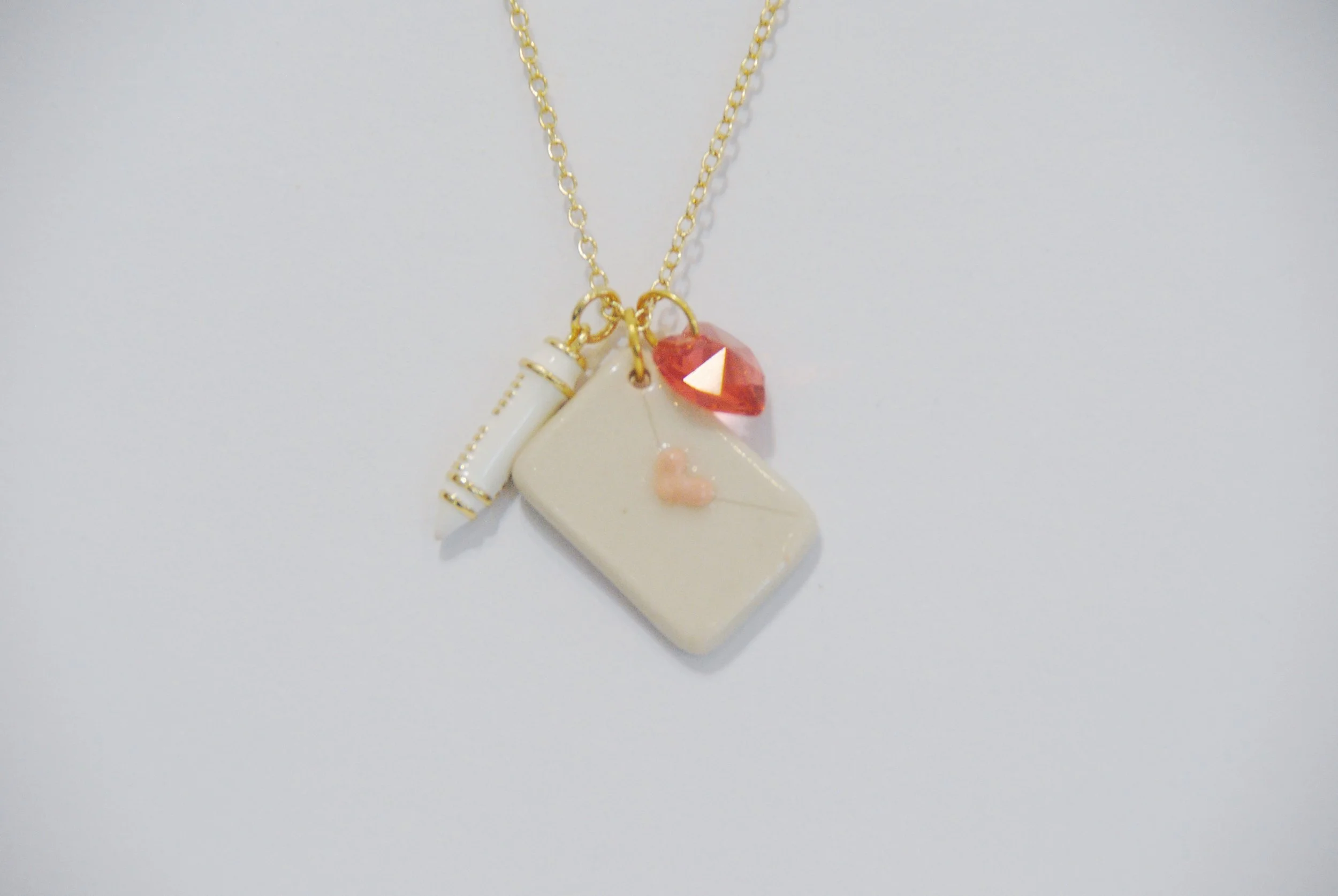 Gold chain necklace with charms including a white lipstick, a white card with a pink heart, a pink gemstone, and a small white scroll.