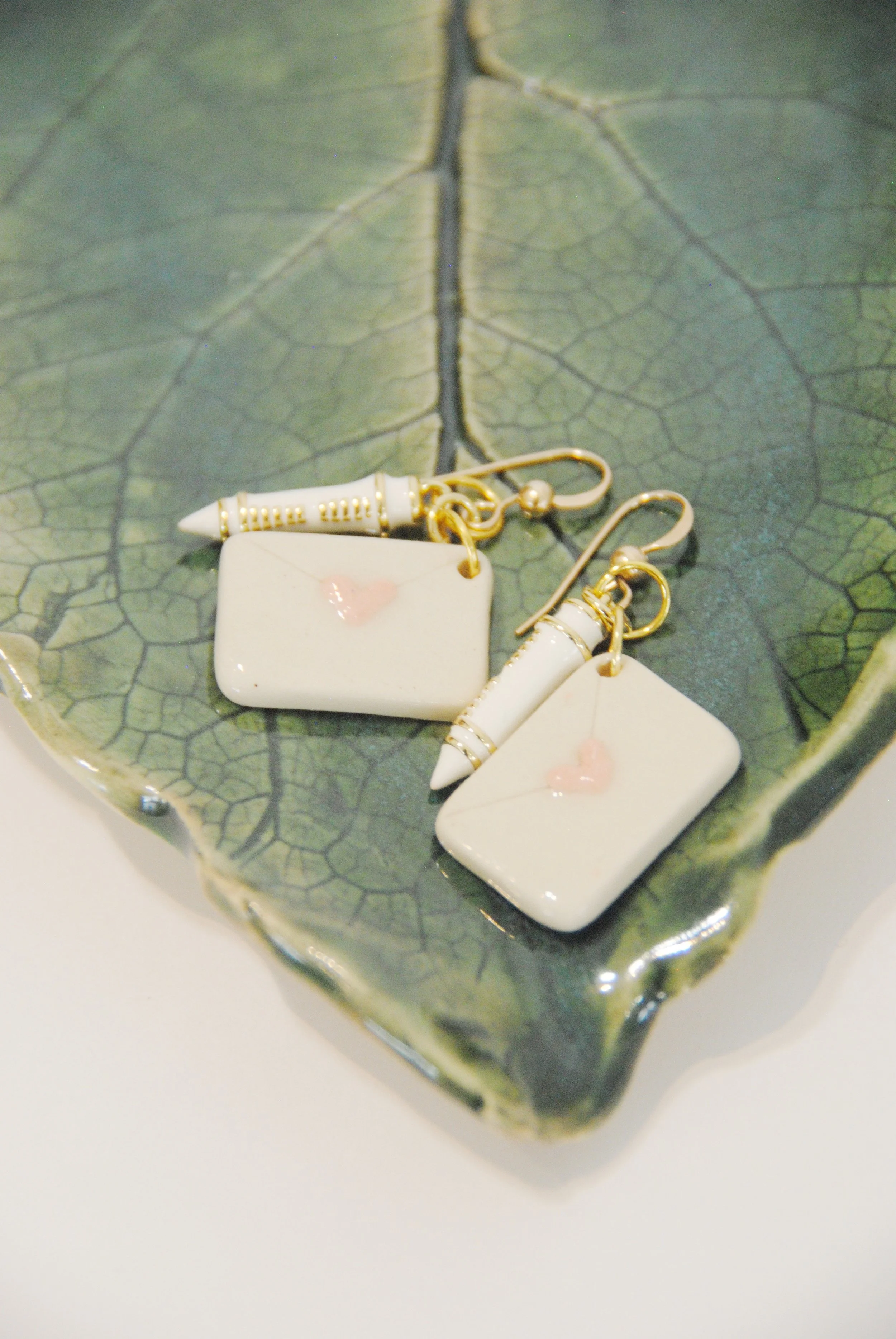 Cream-colored earrings with small pink hearts and decorative white and gold pens, placed on a green leaf-shaped dish.