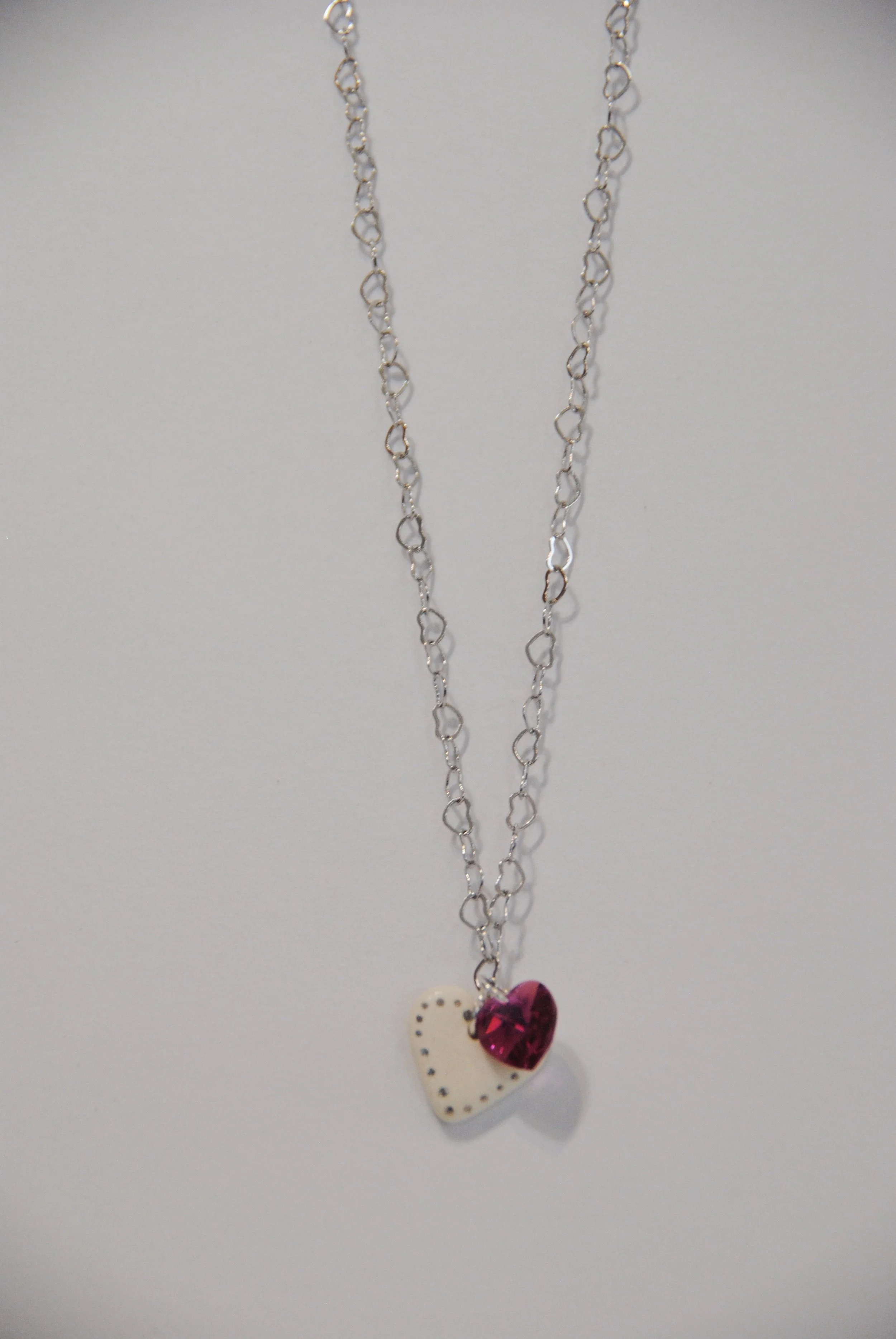 Silver necklace with two heart-shaped pendants, one white with black dotted border and one pink gem, hanging against a light background.