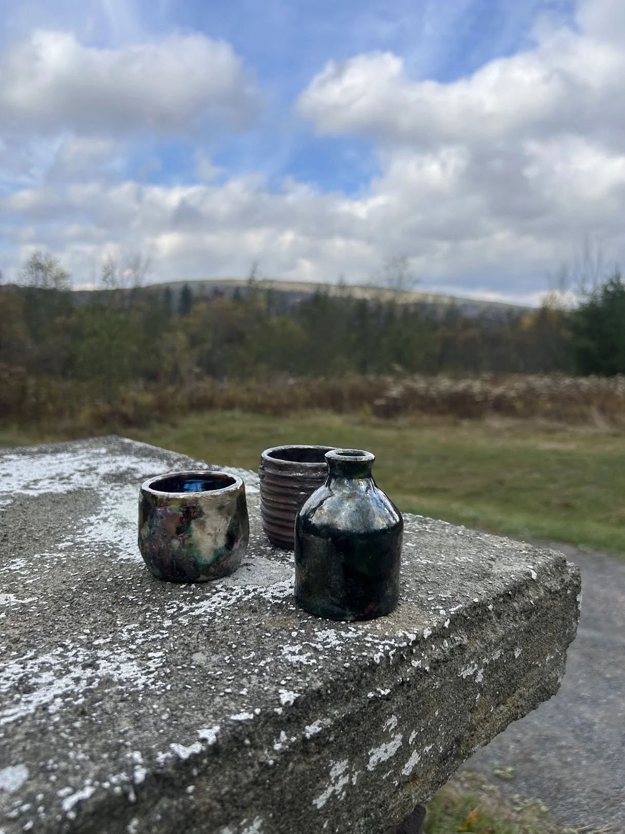 Three small ceramic pots or vases placed on a weathered concrete ledge outdoors, with a natural landscape including grass, trees, and a cloudy sky in the background.