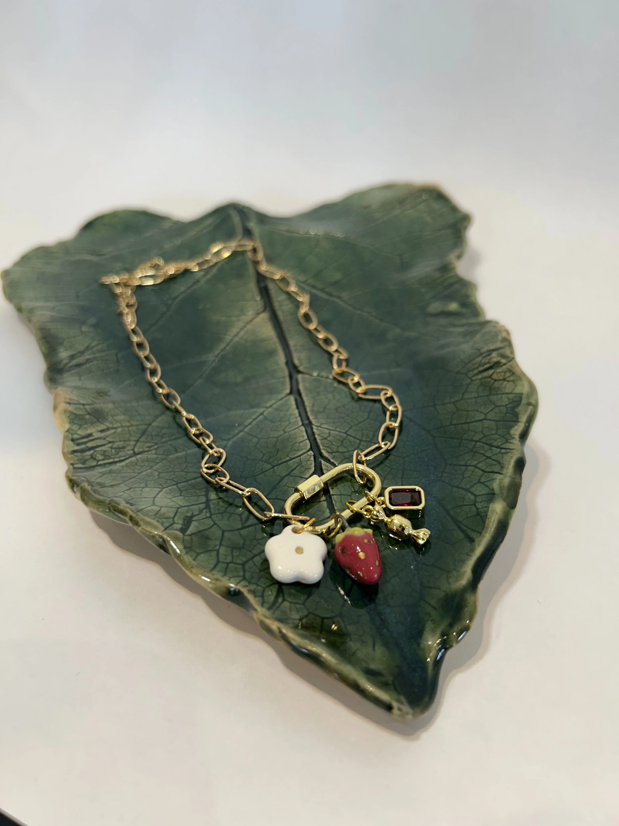 Gold chain necklace with charms of a white flower, a strawberry, a black square gem, and a gold bell, arranged on a ceramic leaf-shaped dish.