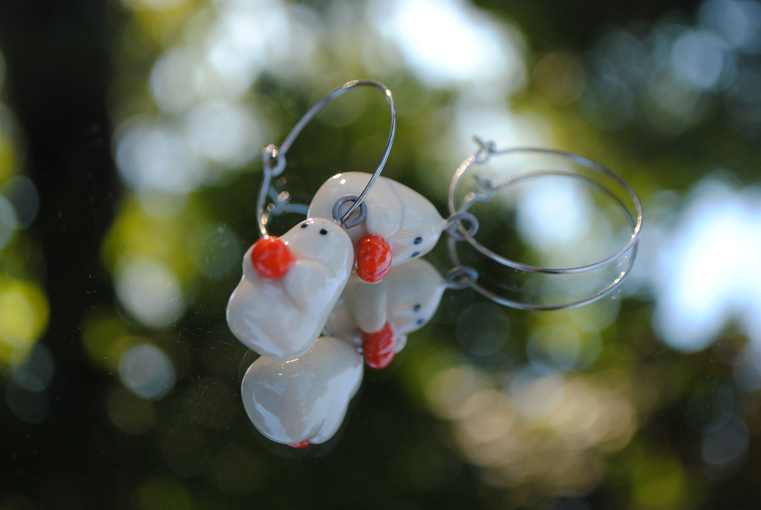 Close-up of ceramic earrings shaped like white mice with red noses and black eyes, hanging from thin metal hooks, with a blurred background of green and white bokeh.