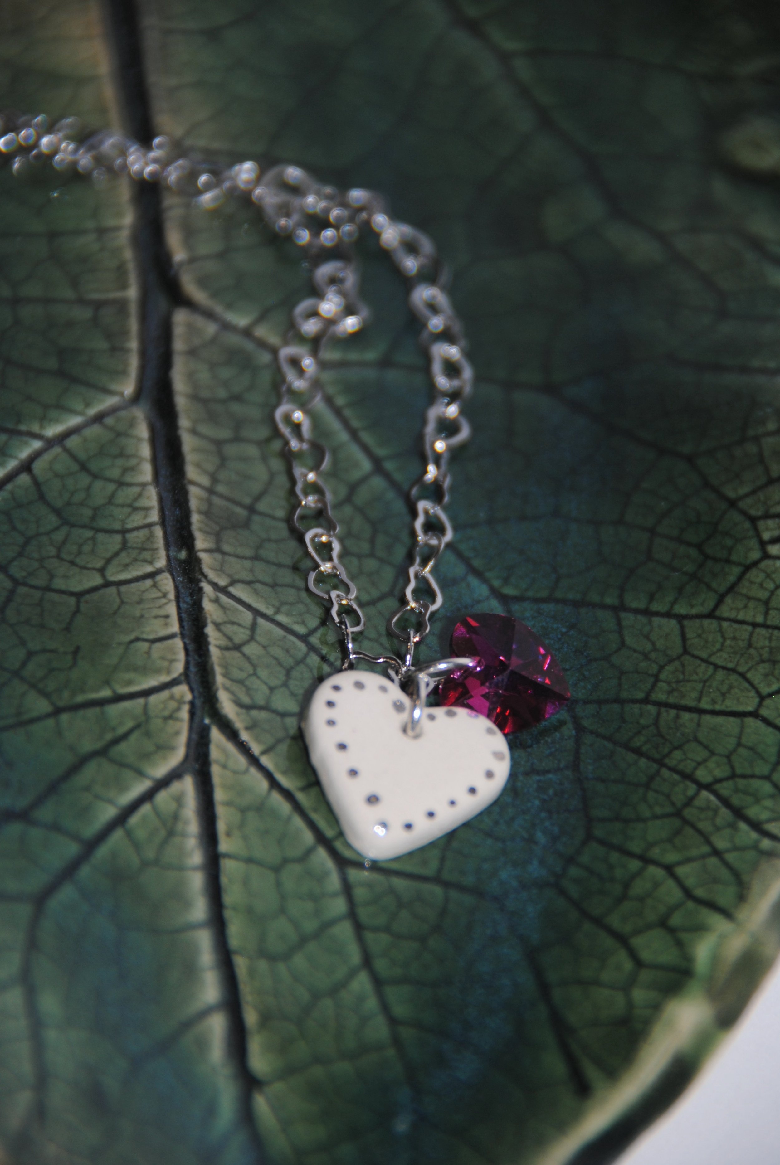 A silver necklace with a white heart-shaped pendant and a small red gemstone charm, placed on a textured green leaf.