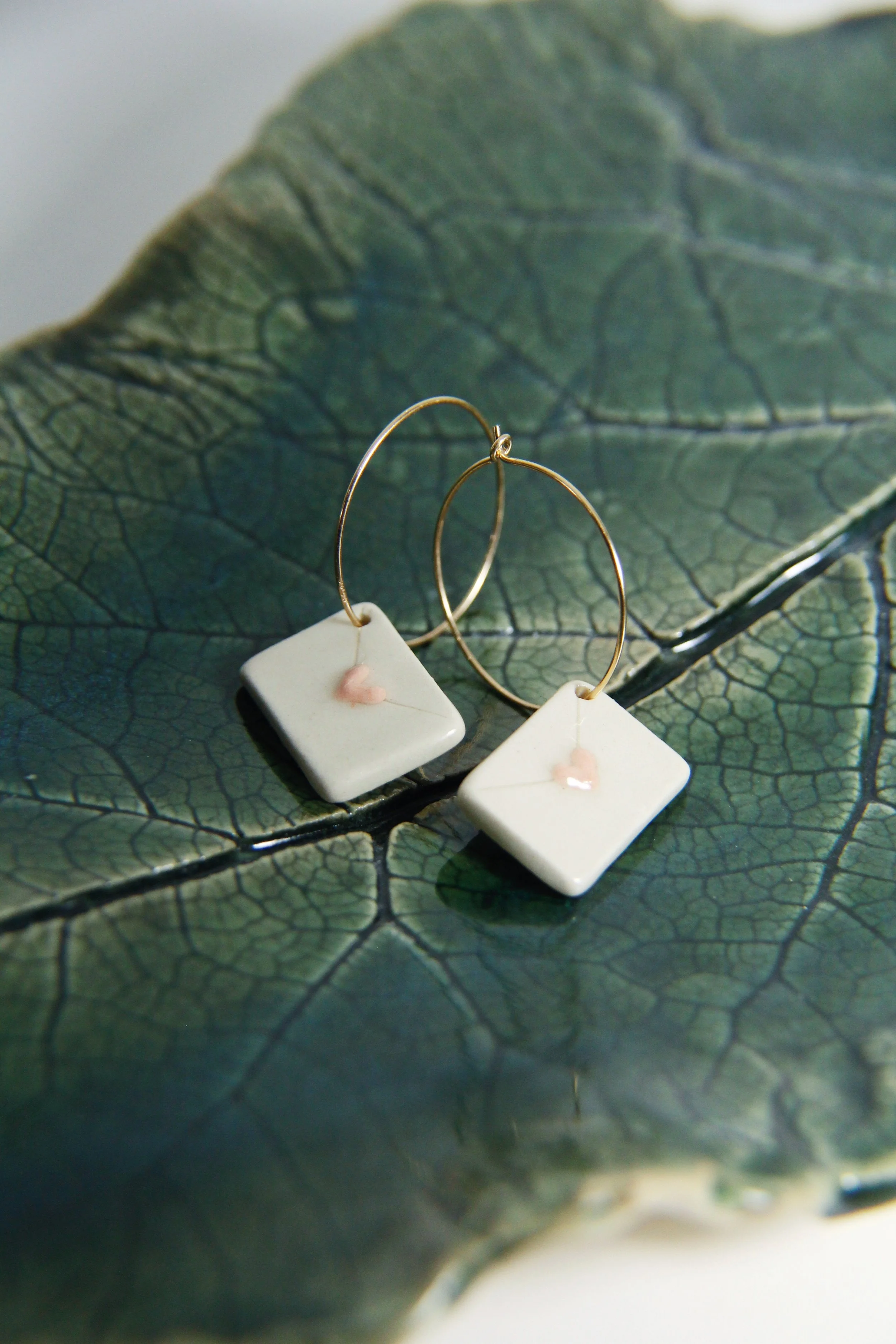 A pair of white ceramic square earrings with small pink heart details, placed on a large green leaf with visible veins.