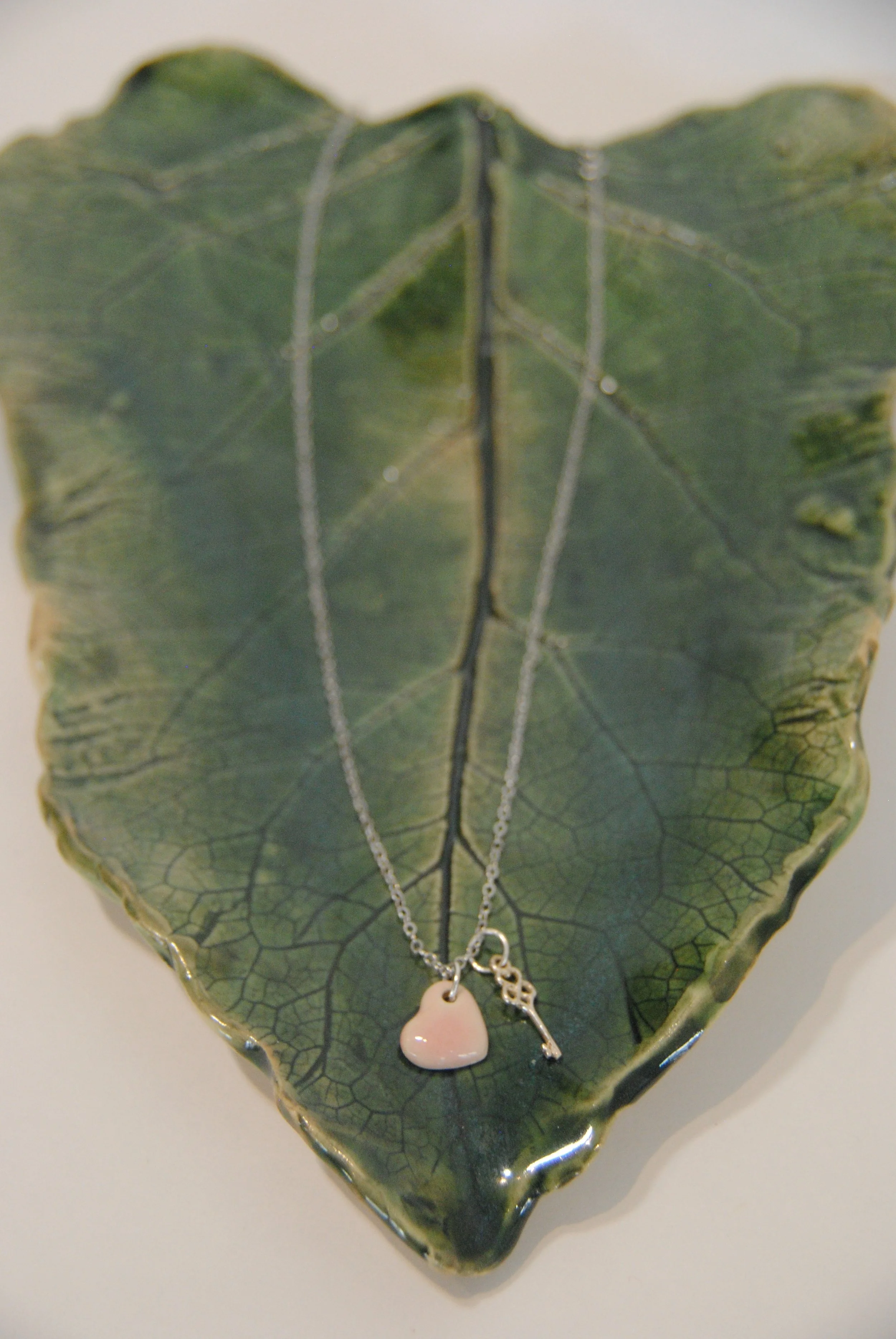 A silver necklace with a heart-shaped pendant and a small key hanging from a leaf-shaped dish.