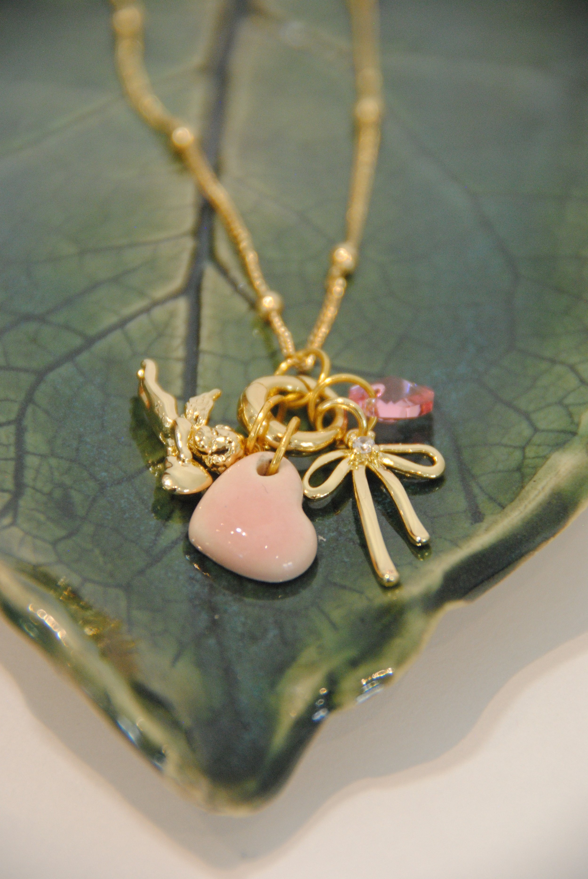 Gold necklaces with charms, including a pink heart, a bow, a pink gemstone, and a bird, on a green leaf-shaped dish.