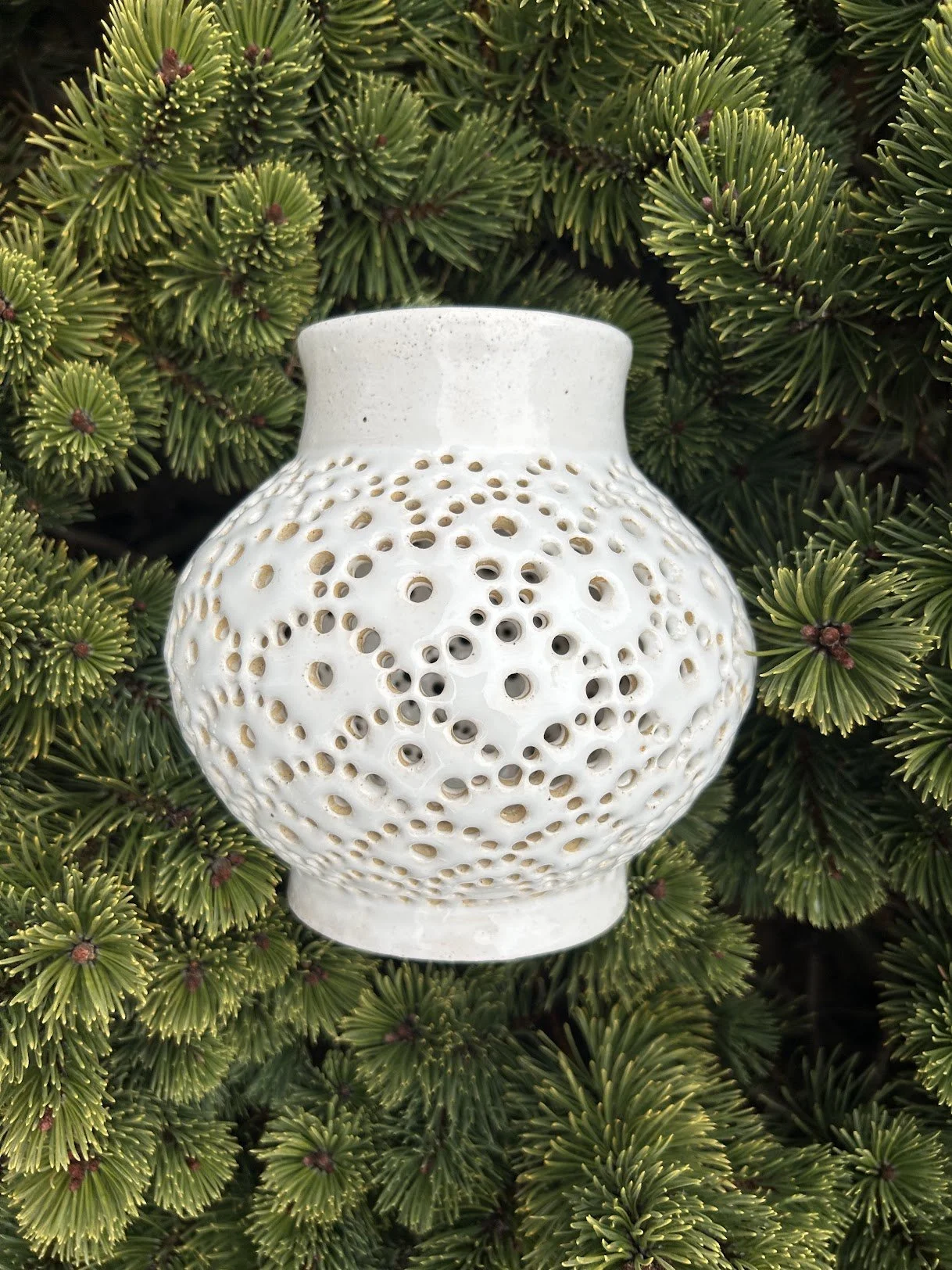 White ceramic vase with numerous circular holes in a pattern, set against a background of green pine branches.