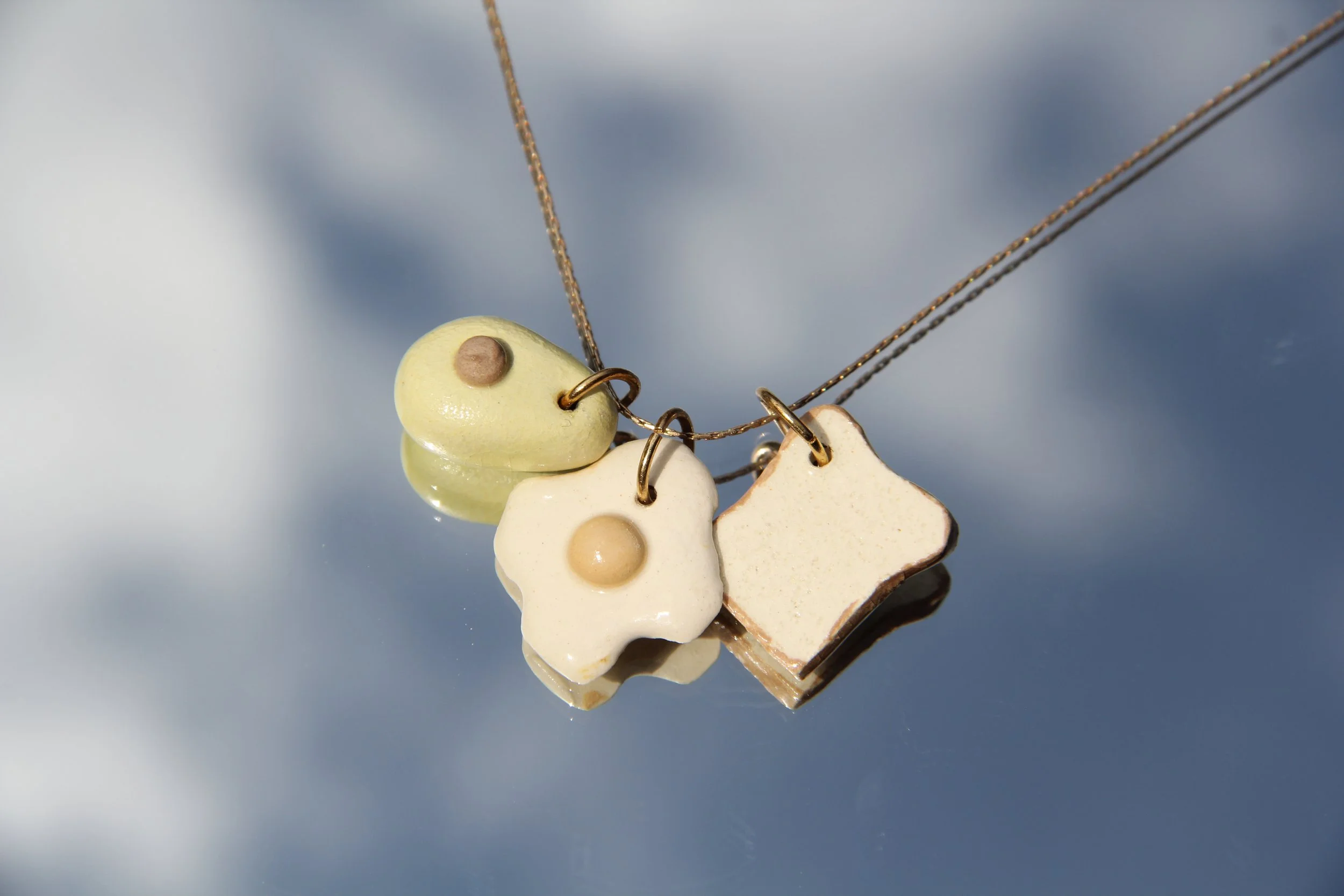 Three ceramic charms shaped like a chicken leg, fried egg, and star, hanging on delicate gold chains against a shiny reflective surface.