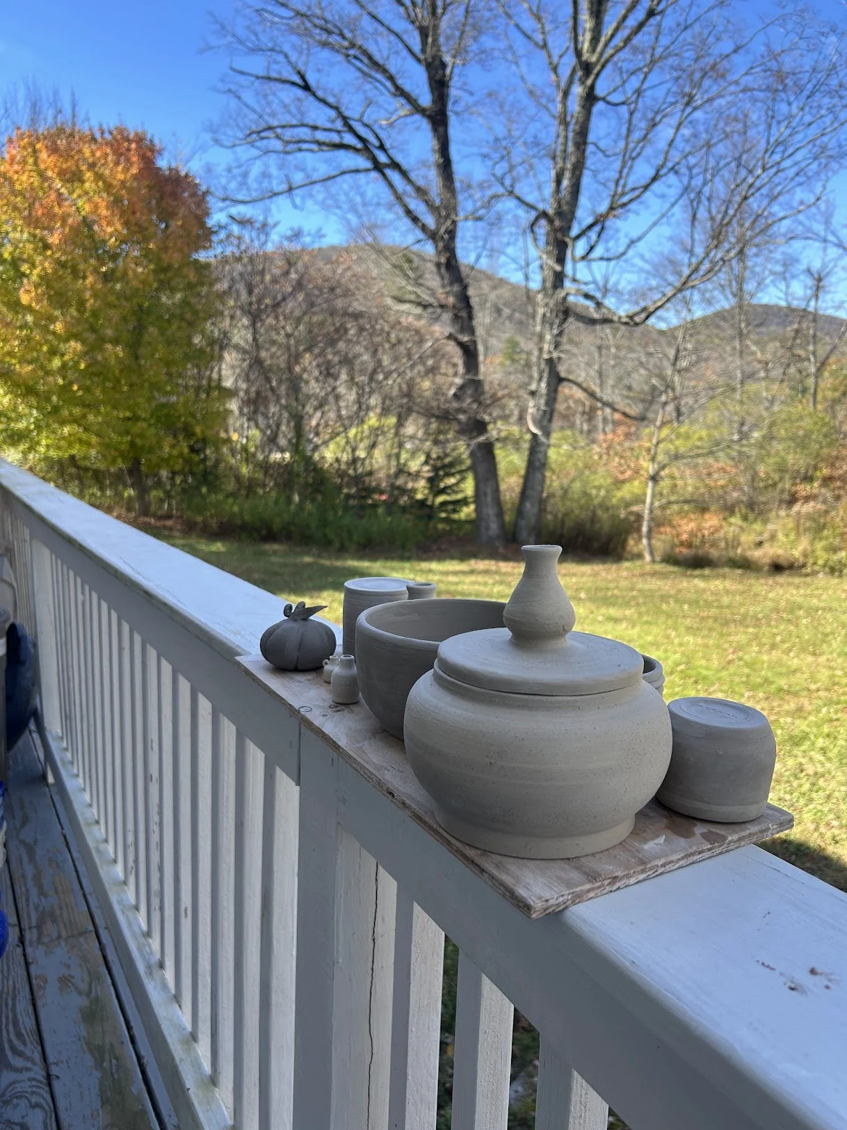 Several unglazed pottery vases and containers on a wooden board resting on a white outdoor railing, with trees and mountainous landscape in the background on a clear day.