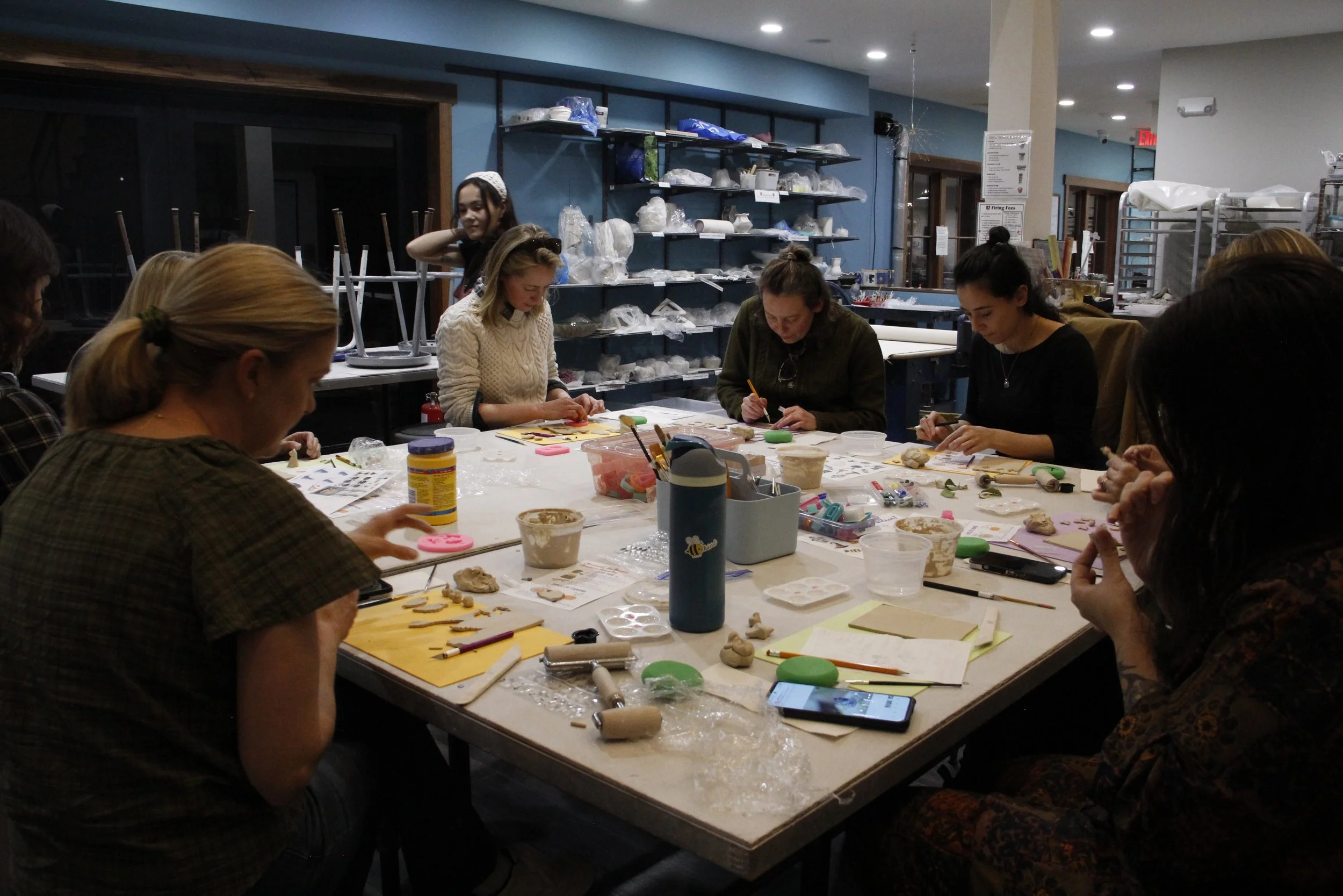Group of women participating in a craft workshop at a large table filled with craft supplies and tools, in a well-lit indoor space.