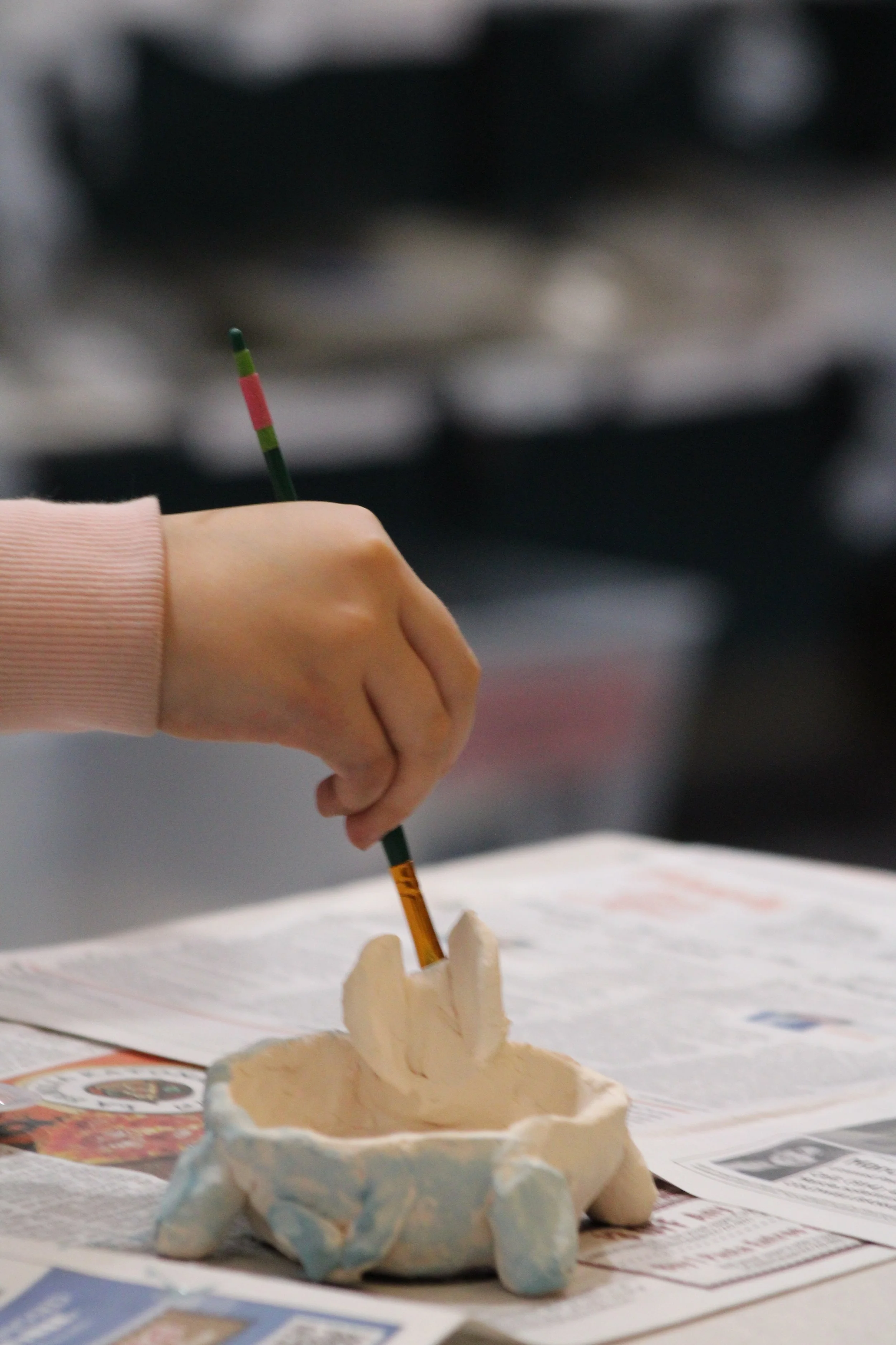 A person is painting a ceramic object with a small brush in a workspace. The ceramic piece resembles a small dish or sculpture.