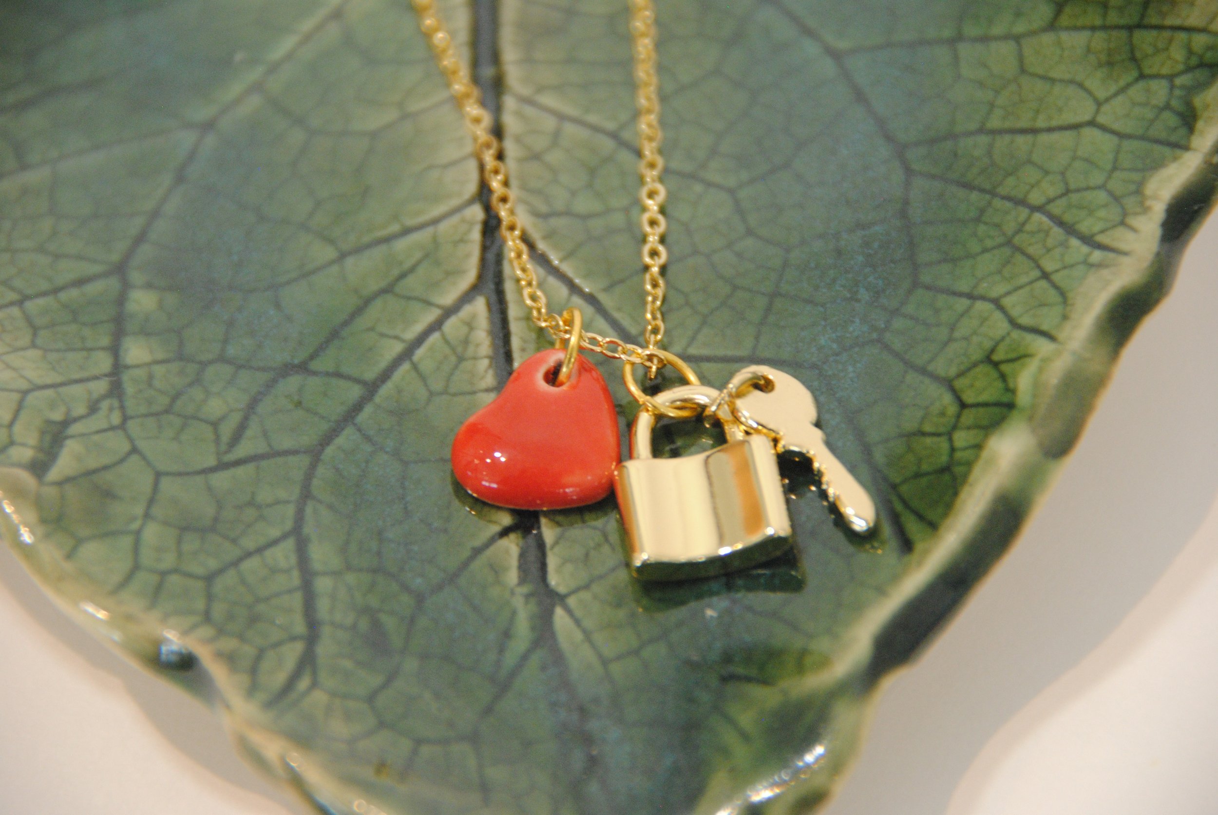 Gold necklace with three charms: a red heart, a gold rectangular charm, and a small gold key, resting on a green leaf-shaped ceramic dish.