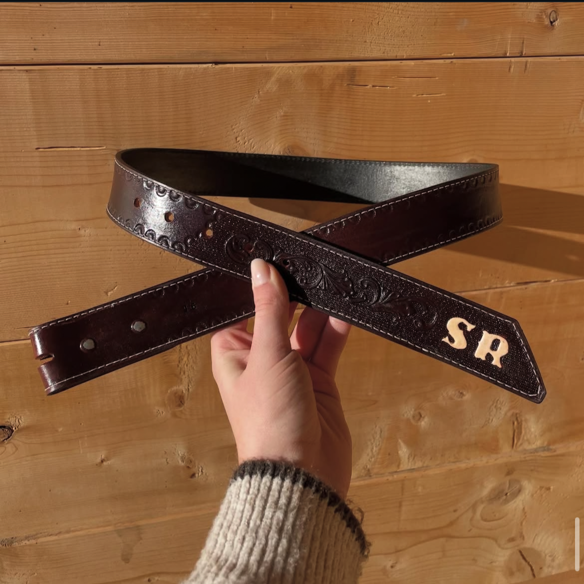 A person holding a dark brown leather belt with decorative embossing and the initials 'SR' in white on the end, against a wooden background.