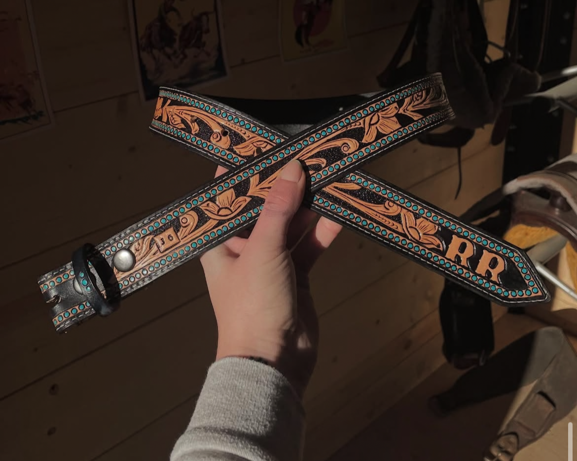 A hand holding a decorative leather belt with intricate black, brown, and turquoise patterns, including floral designs and the letters 'J' and 'R'.