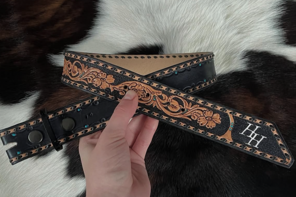 Hand holding a decorative black leather belt with floral designs and turquoise accents over a black and white cowhide background.
