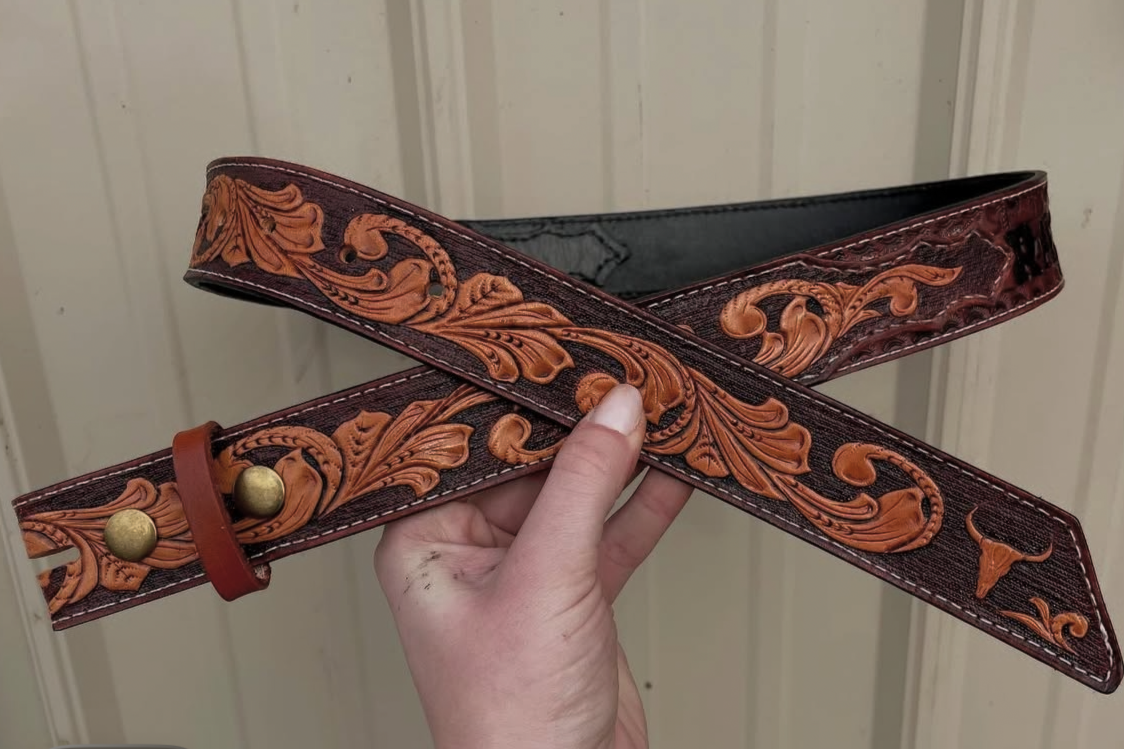 A hand holding a brown leather belt with ornate, embossed floral and bull head designs.