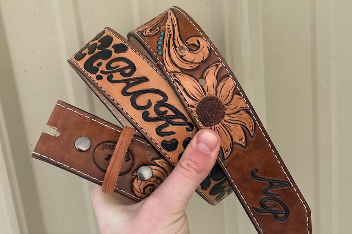 Hand holding multiple brown leather belts with engraved patterns and text, some with metal snaps, against a light-colored wood background.