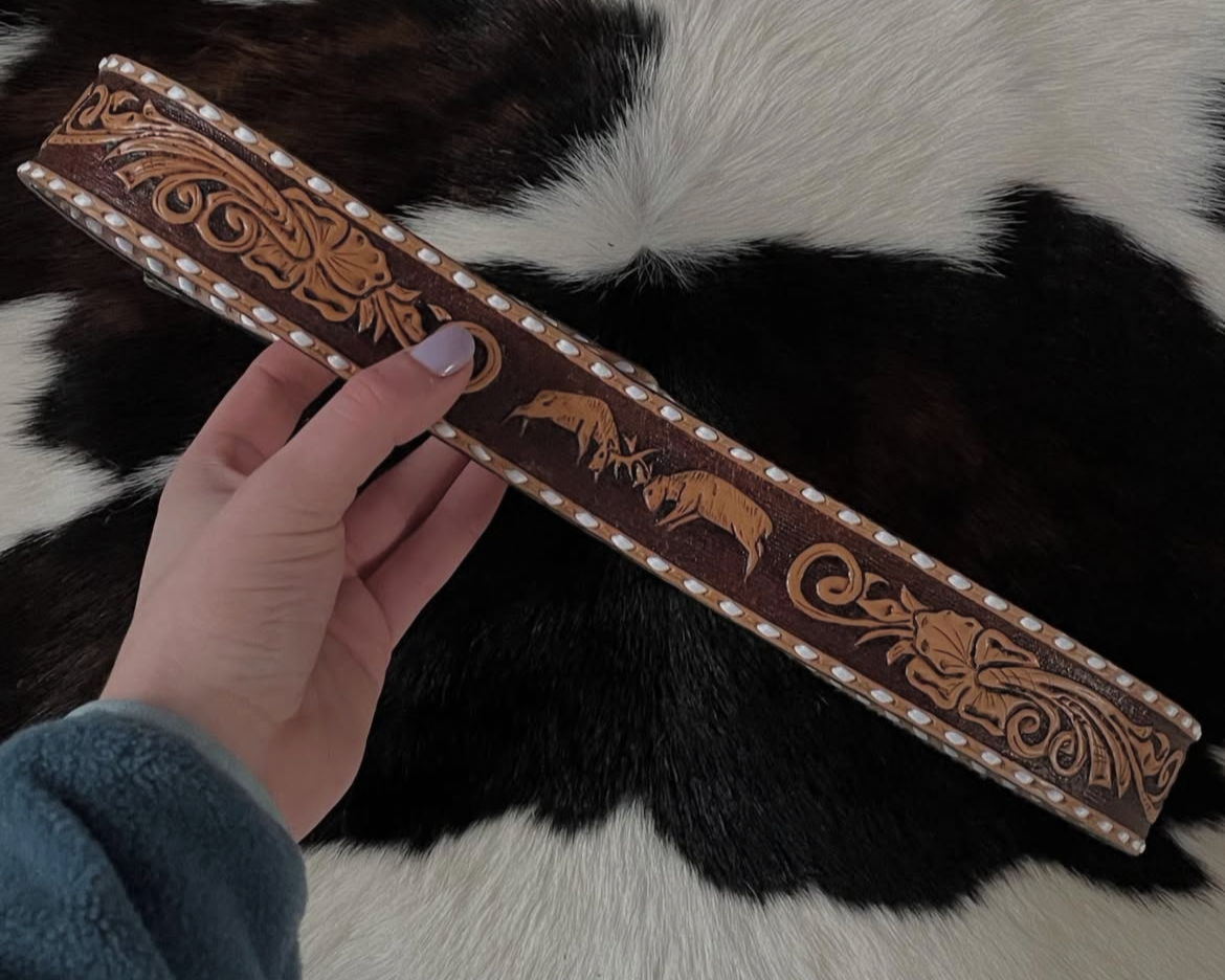 A person holding a decorative leather belt with carved designs, laid on a black and white cowhide rug.