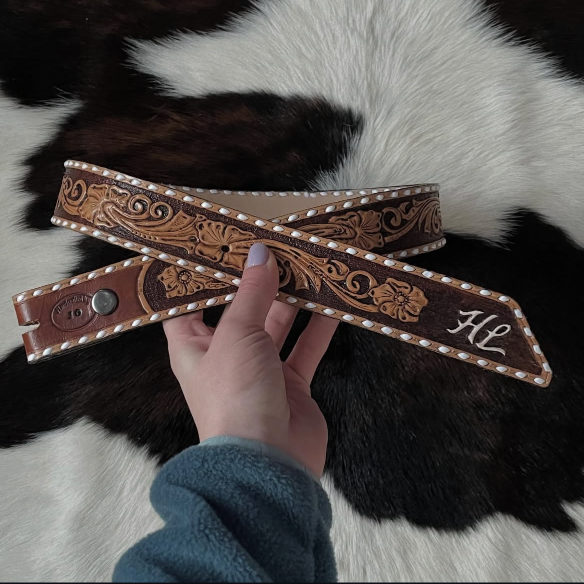 A person holding a decorative leather belt with intricate floral carvings and a metal snap, placed on a black and white cowhide rug.