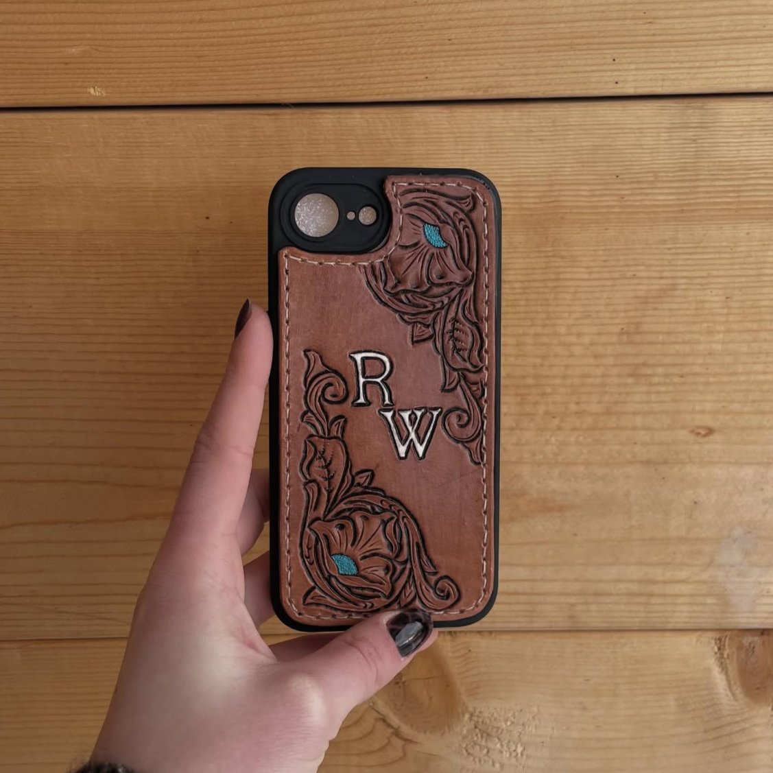 A person's hand holding a smartphone with a leather phone case decorated with intricate floral embroidery and the initials 'R W' on the back, against a wooden background.