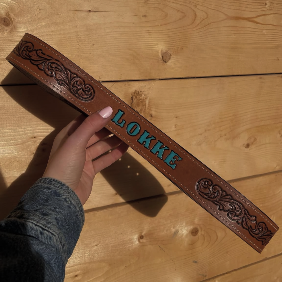 A leather belt with intricate embossed designs and the word "LONKE" in blue letters against a wooden background. A person's hand with light-colored nail polish holds the belt.