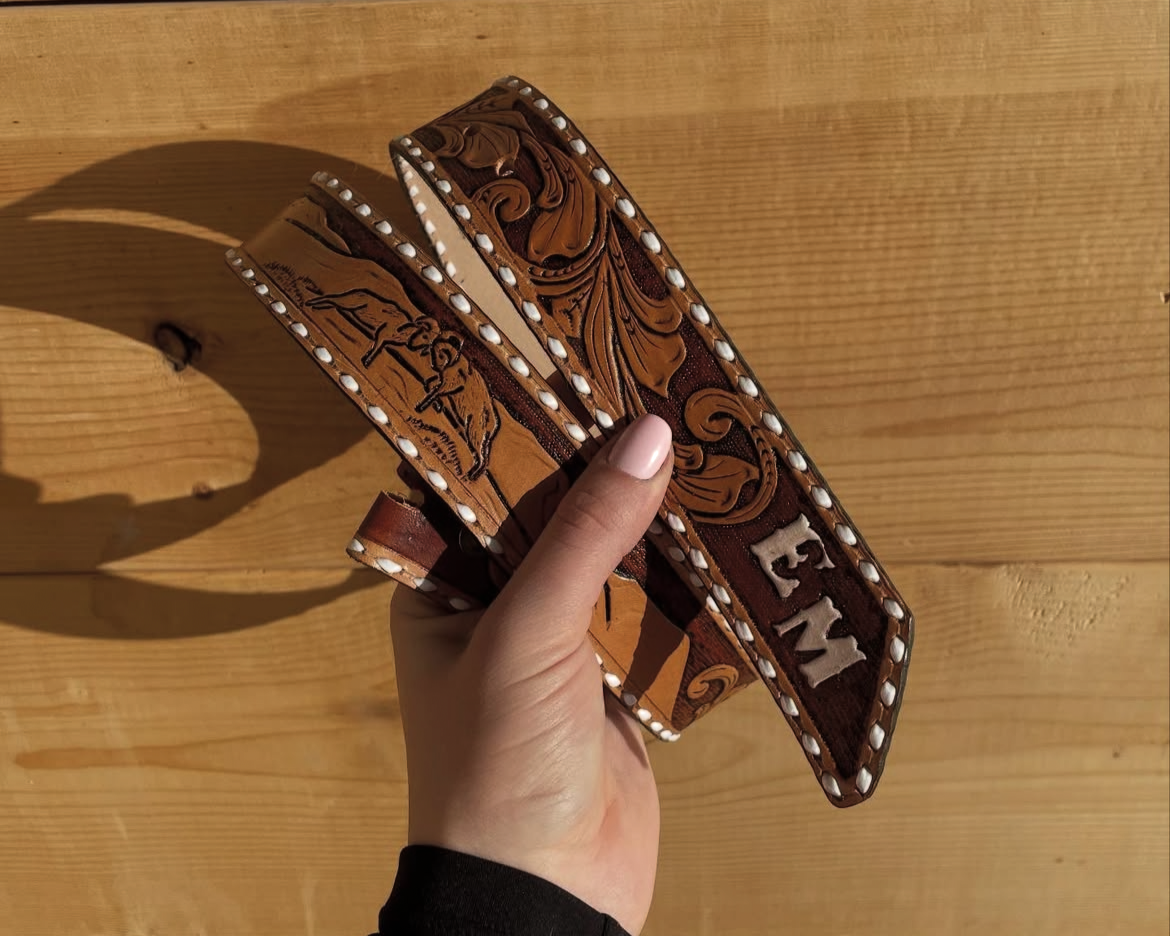 A hand holding a decorated western-style leather belt with silver studs and detailed embossing, against a wooden background.