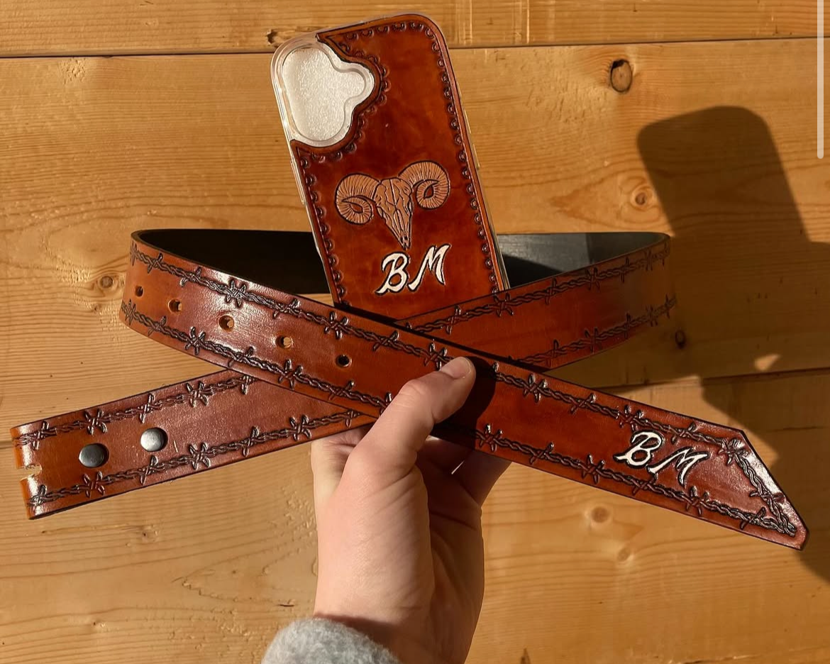 A leather belt with decorative stitching and metal snaps, and a leather holster with a ram skull design and initials 'BM' on a wooden surface.