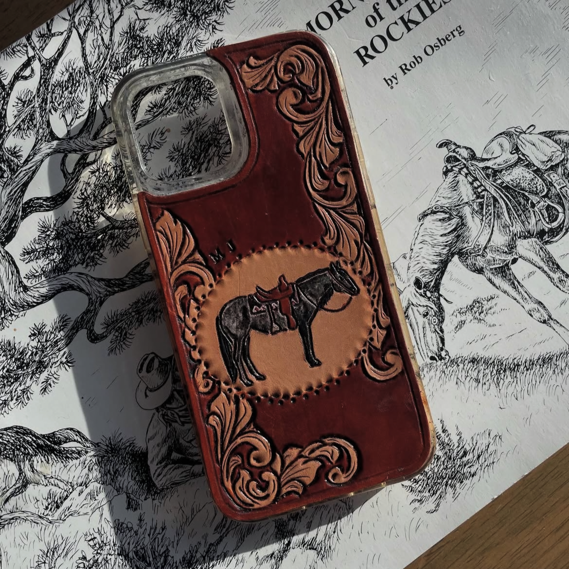 Leather phone case with a cowboy horse design resting on a black-and-white drawing of a cowboy riding a bucking horse.